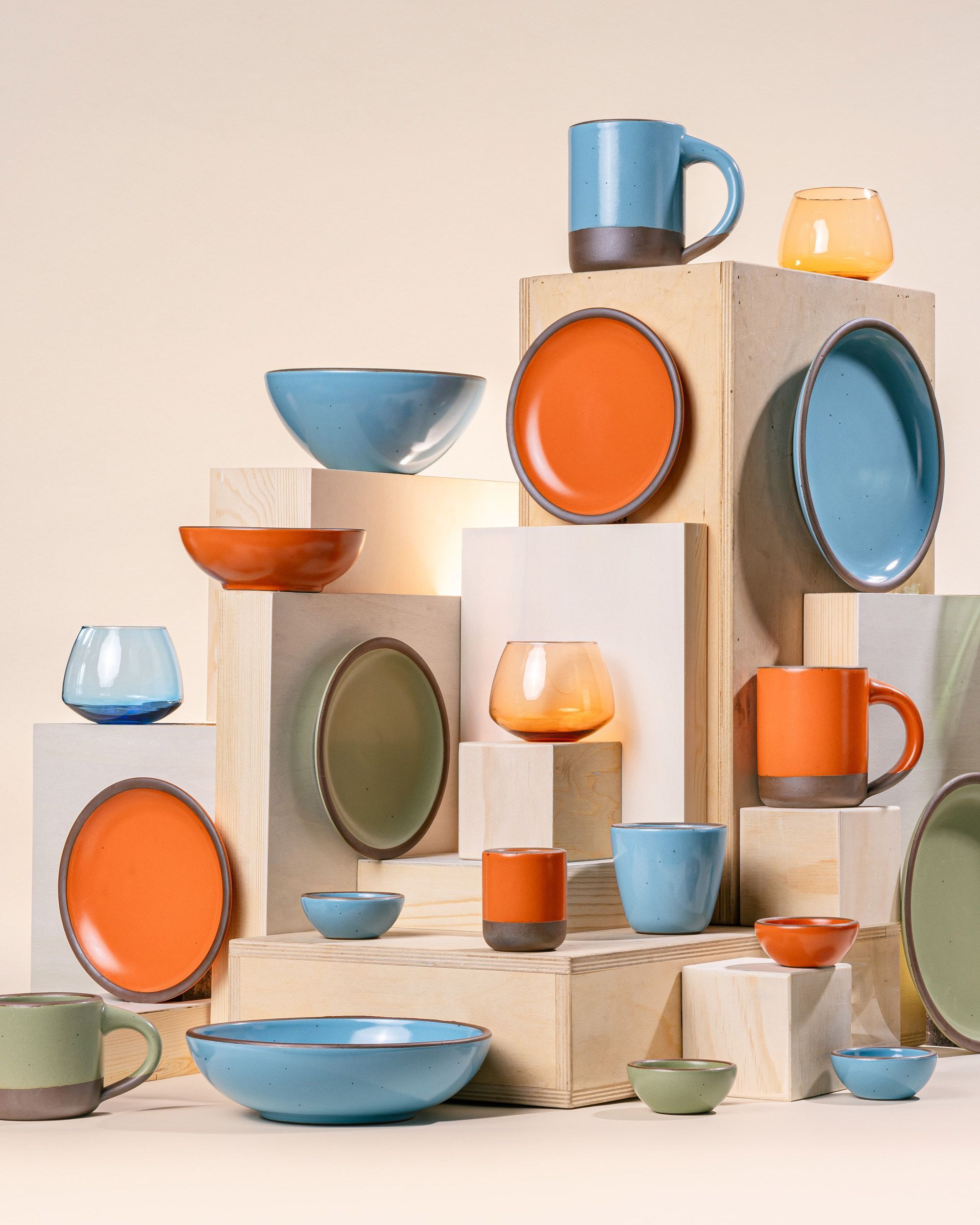 An artful arrangement of ceramic dinnerware in bold orange, calming sage, and robin's egg blue colors, arranged at various heights and positions on wooden risers.