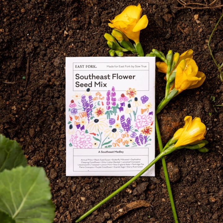A white seed pack that reads "Southeast Flower Seed Mix" with colorful floral stem illustrations. The seed pack is sitting in soil next to some flower stems
