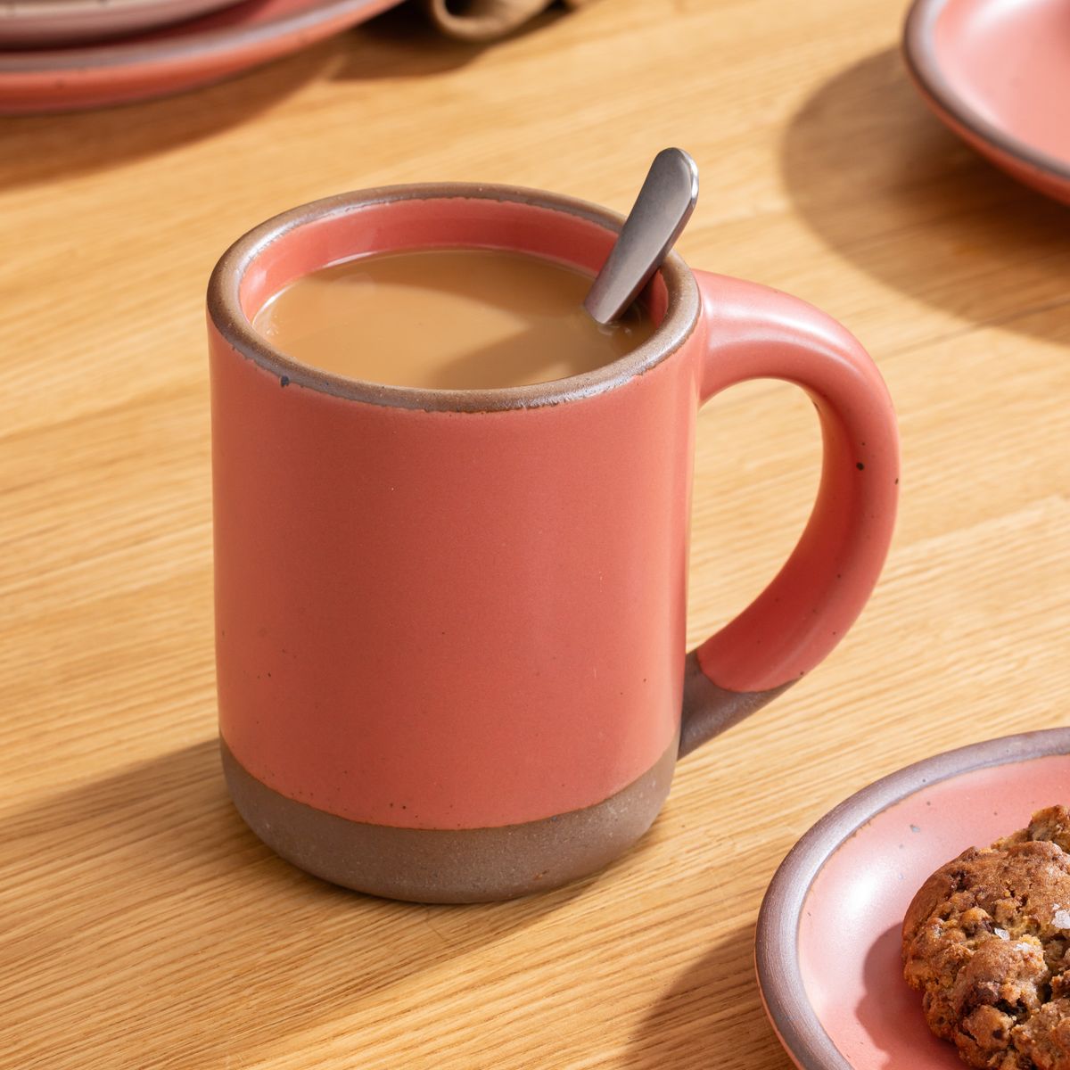 Watermelon pink, big ceramic mug with coffee and spoon, beside a matching plate holding a partially eaten cookie on a wooden table.