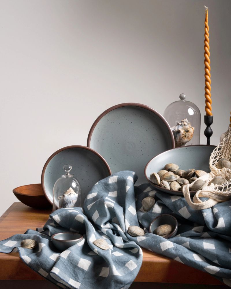 Ceramic plates in a blue-grey color artfully arranged on a table with blue-grey crosshatch fabric, shells, bell jars, and a taper candle.