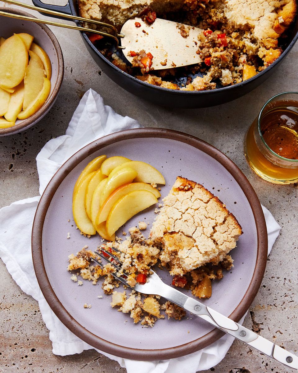 A shepherd's pie and pan-friend apples with a fork on a pastel purple ceramic plate, surrounded by a drink, the cast-iron pie pan, and a bowl of apples.