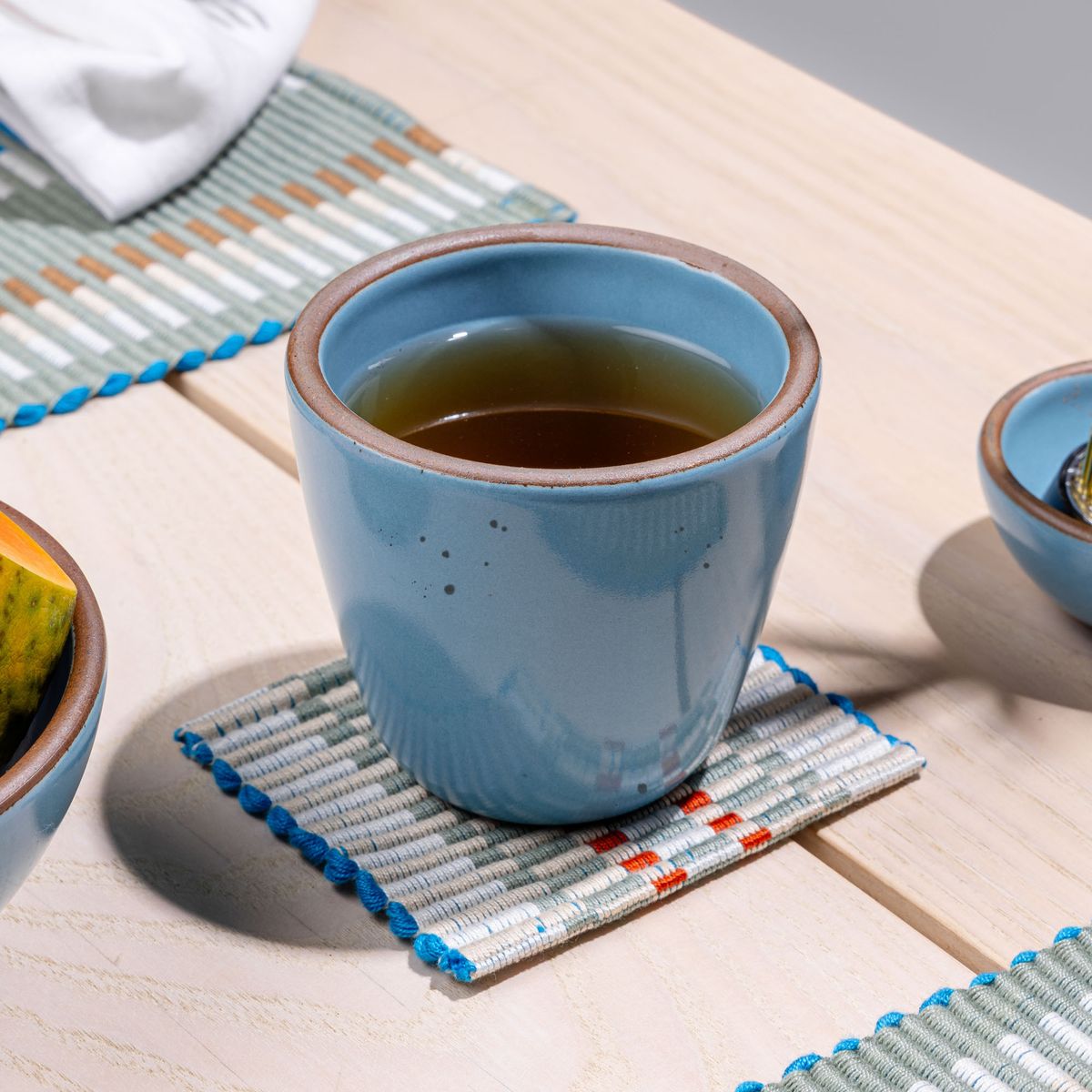 A short cup that tapers out to get wider at the top in a robin's egg blue color featuring iron speckles, sits on a woven coaster on a table.
