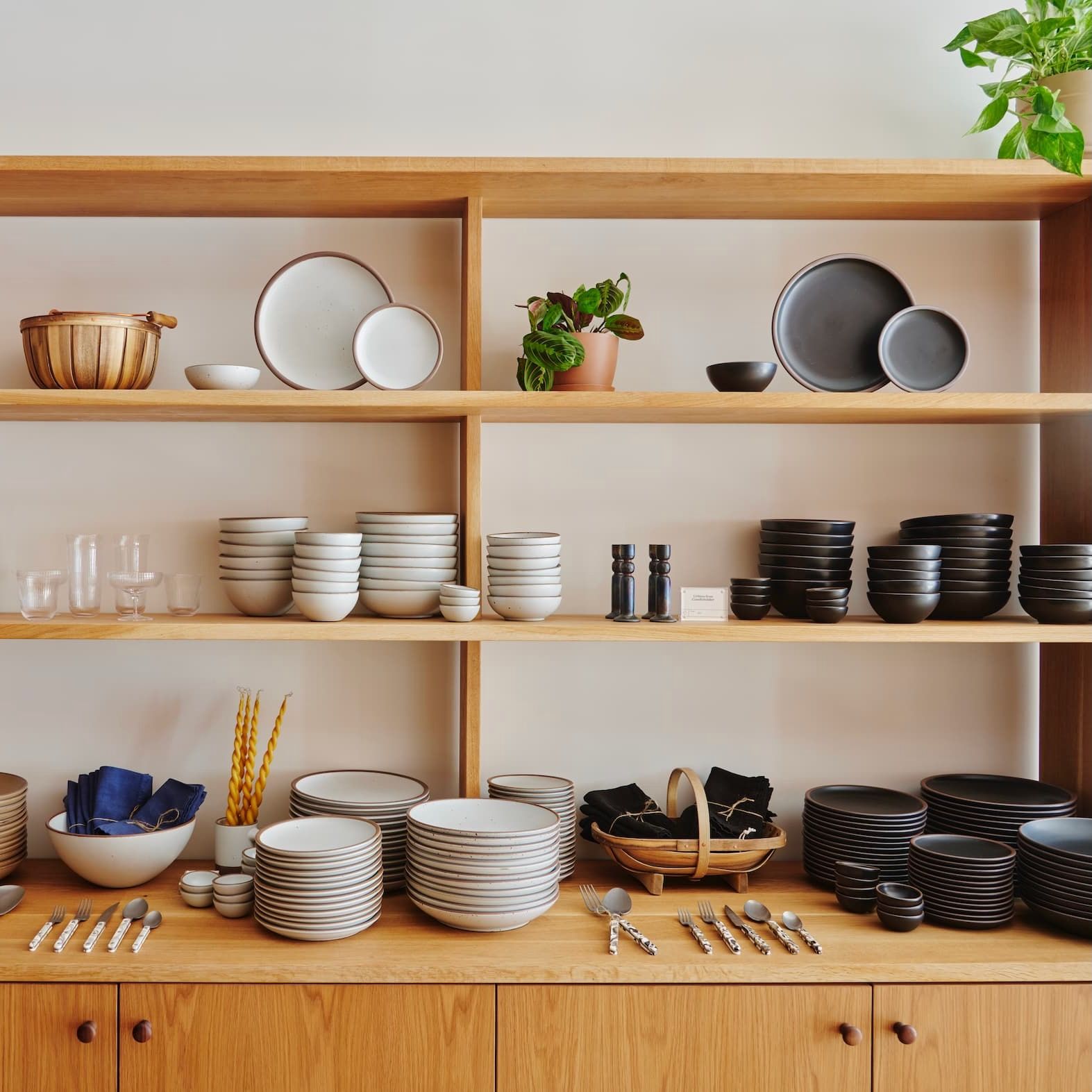 Warm wood shelves highlight neutral tan, white and black ceramic dinnerware with flatware, napkins, candles and more merchandised as well.