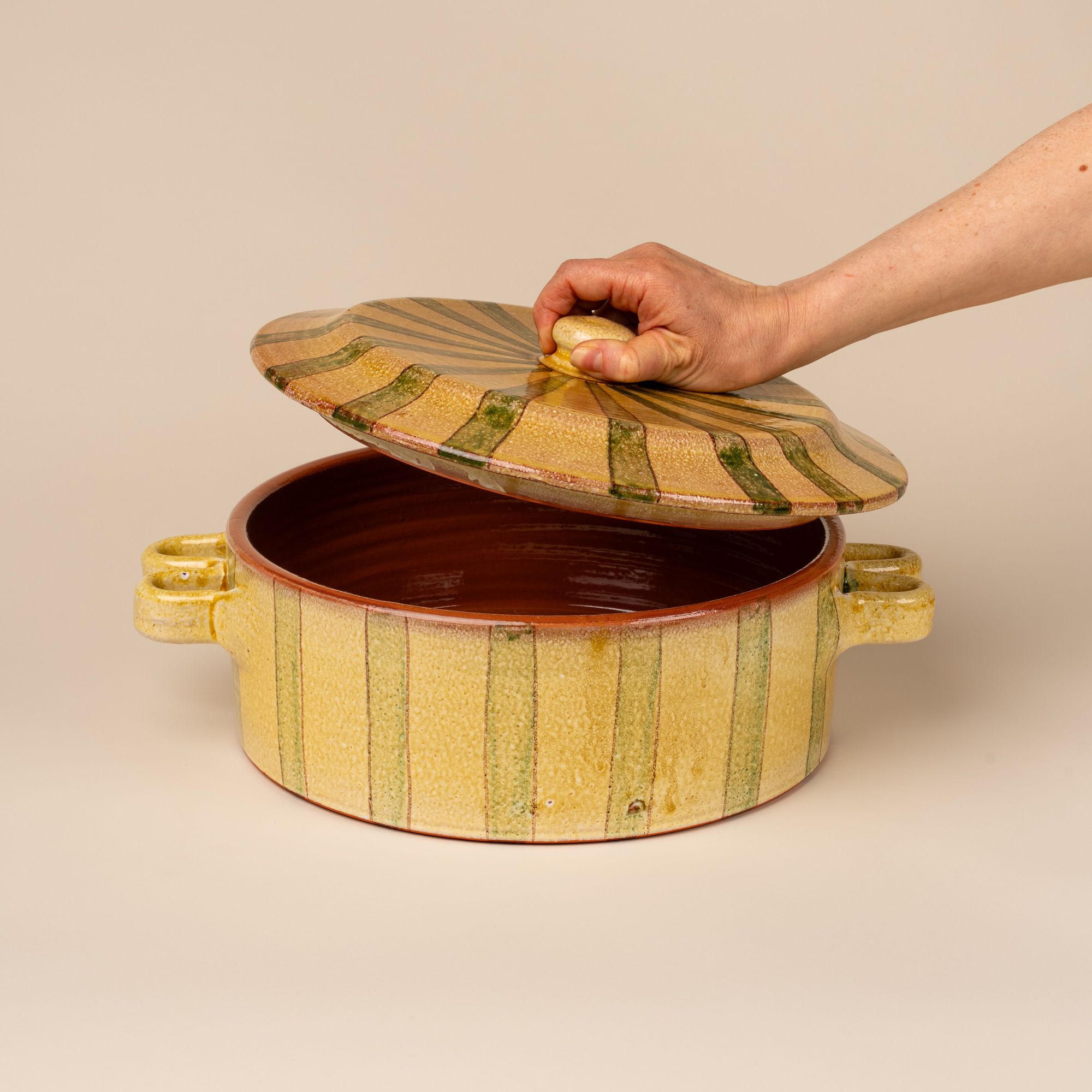 A hand holds the top of a large terracotta casserole dish with handles on each side and large lid with knob handle. The pot is striped with green and butter colors.