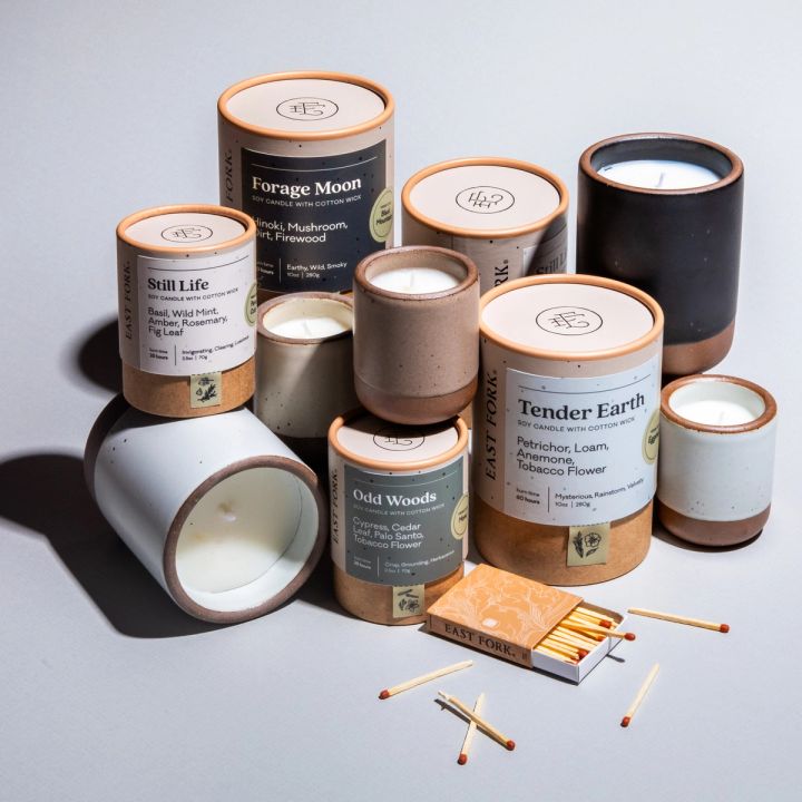 A variety of candles in neutral colors and in ceramic vessels. They're surrounded by cardboard tube branded packaging and a little box of matches.