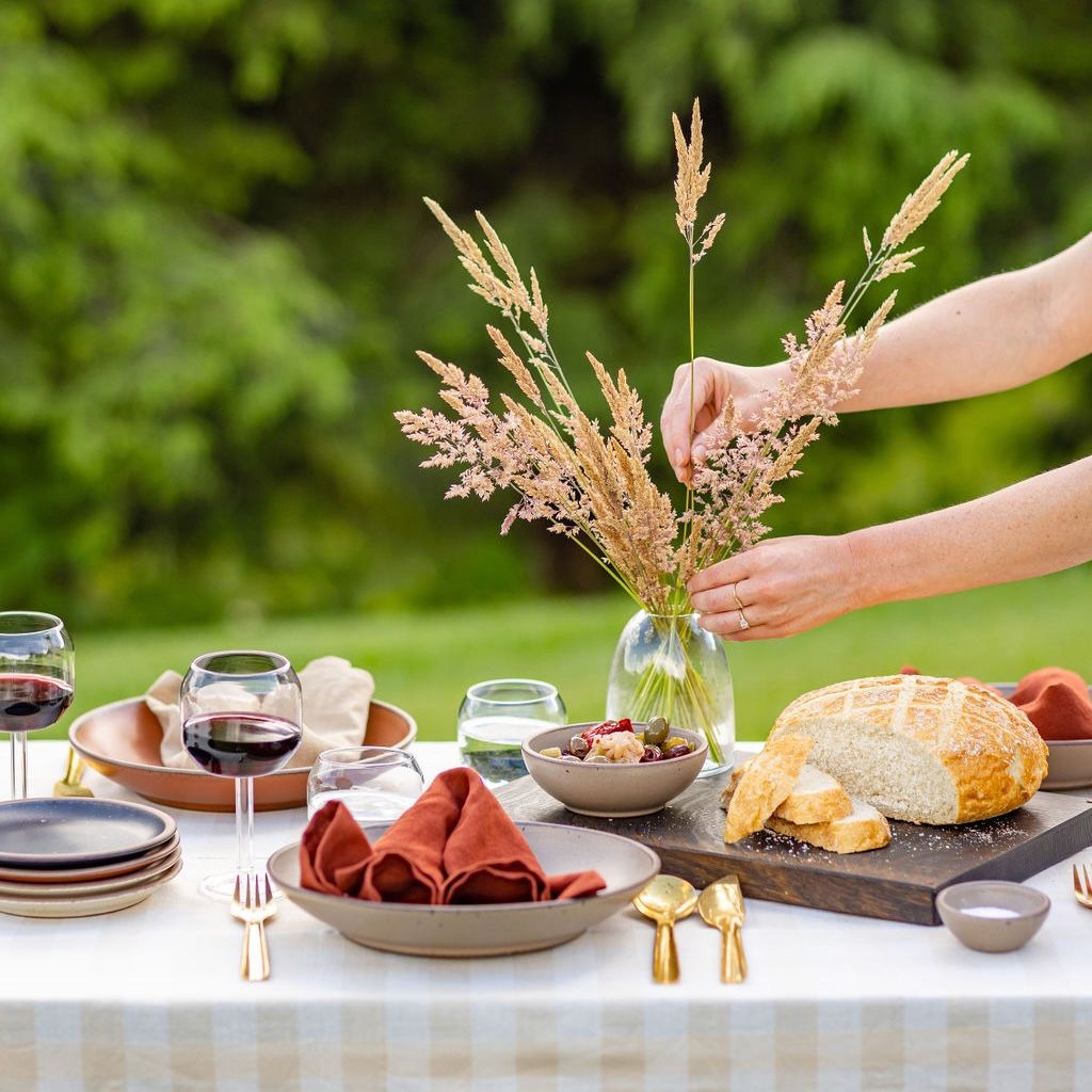 At an outside table, someone is arranging dried florals for a centerpiece. The table is filled with plates, wine glasses, red napkins, loaf of bread, and more.