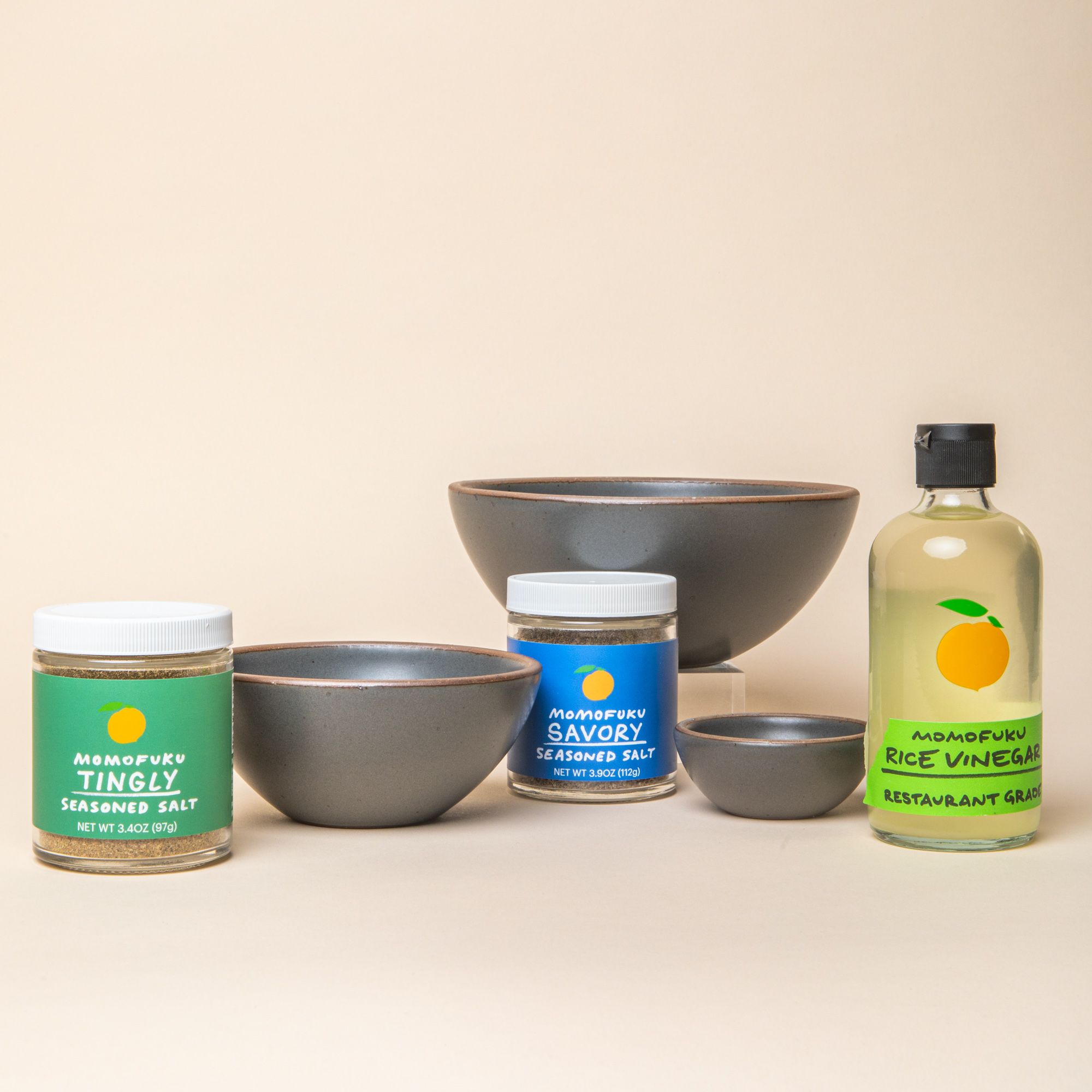 3 Ceramic bowls of varying sizes in a soft cool grey color surrounded by 2 jars of Momofuku seasoned salt and a bottle of rice vinegar.