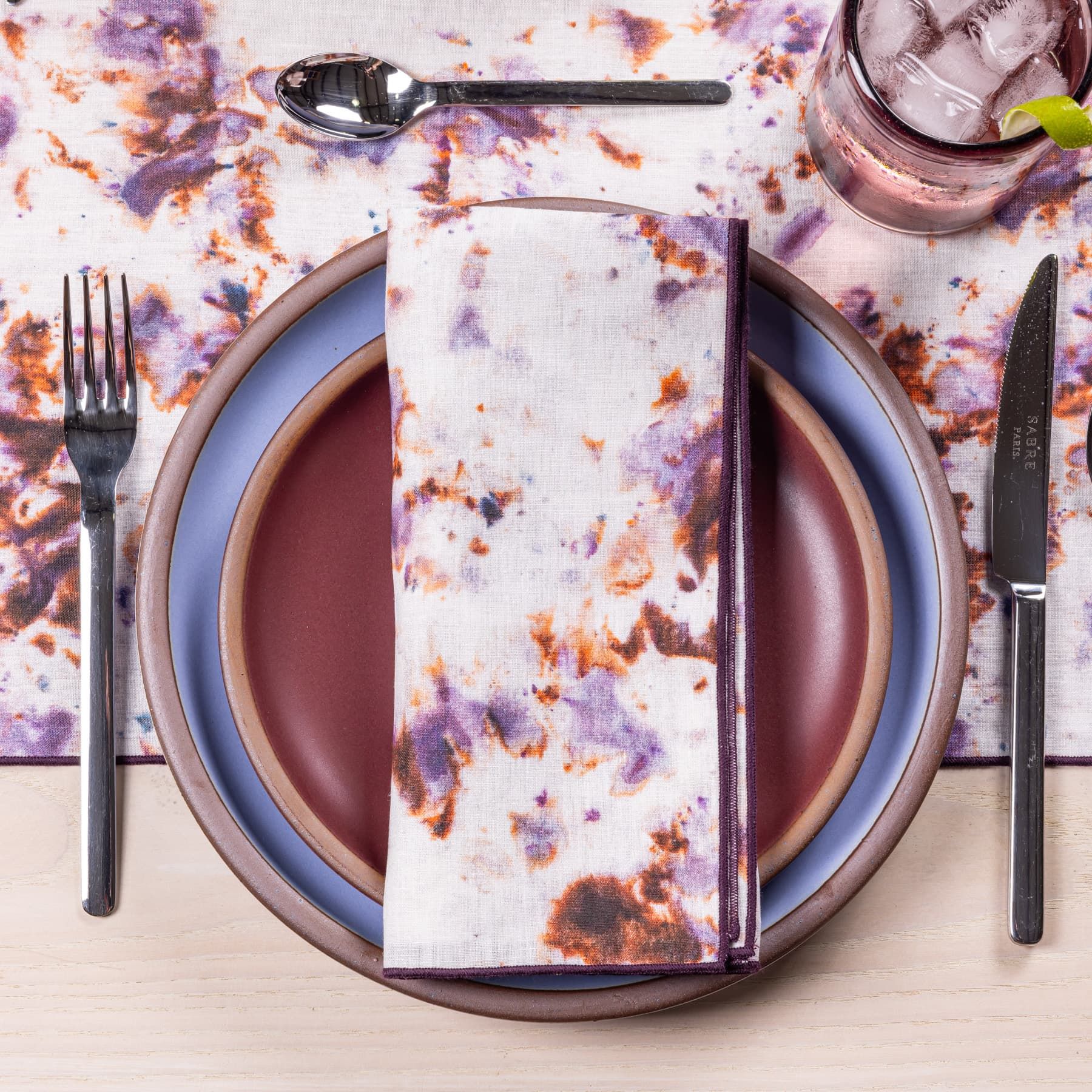 A place setting with periwinkle and plum ceramic plates, a linen napkins in a marbled pattern with warm purple colors, and steel flatware