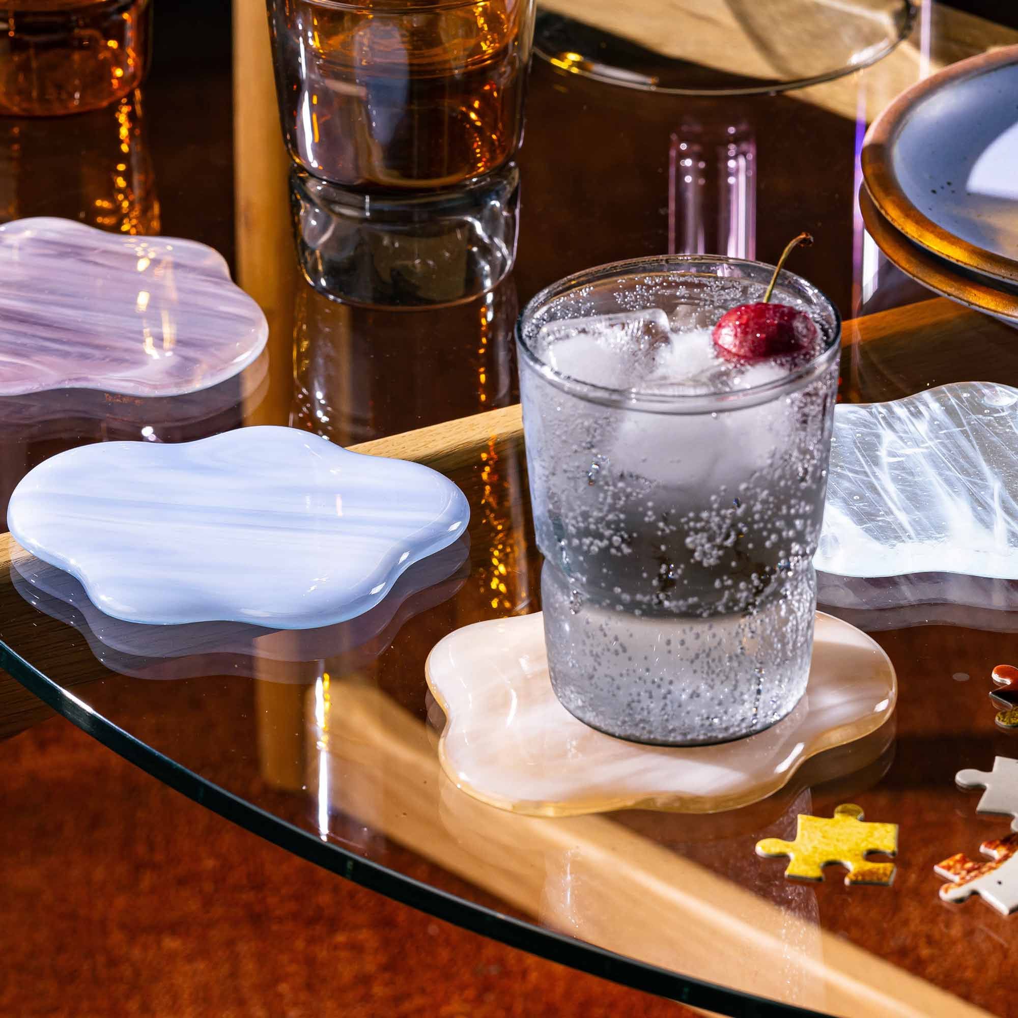 A tumbler glass that widens at the top in a grey color sitting on a coaster that looks like a cloud