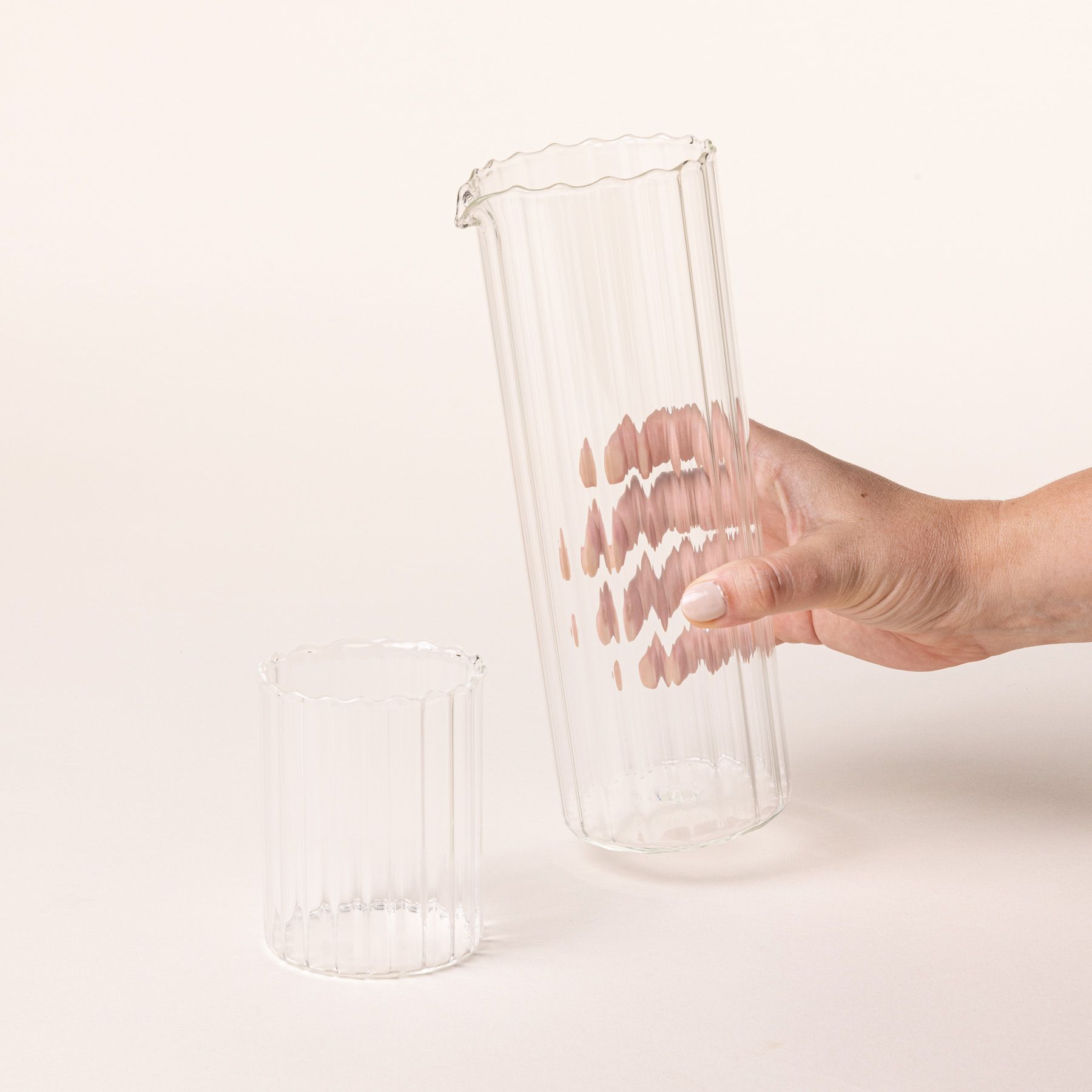 A hand holds a tall cylindrical fluted pitcher with a spout on top and a short tumbler