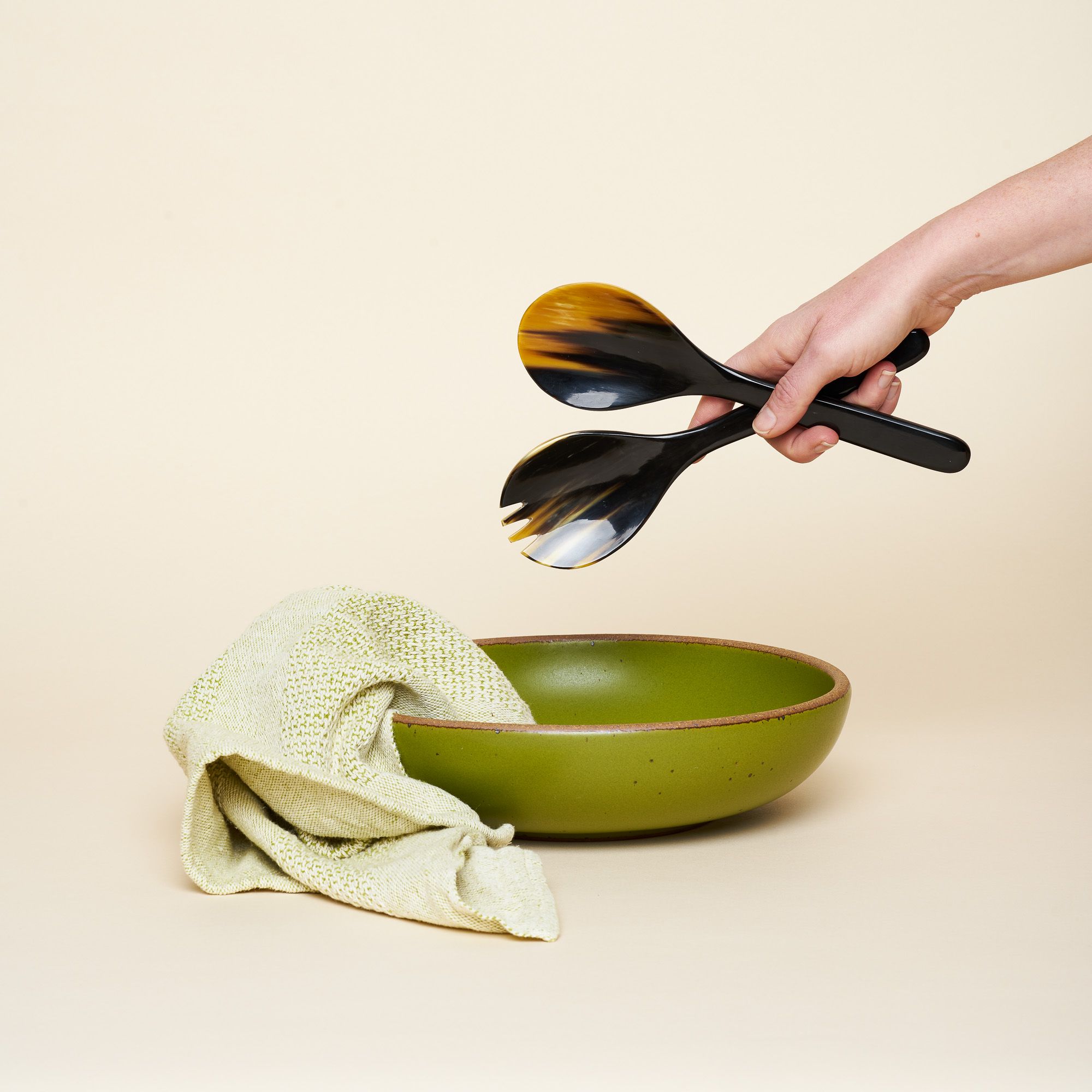 A green shallow but large bowl for serving, with a green tea towel and natural horn salad servers.