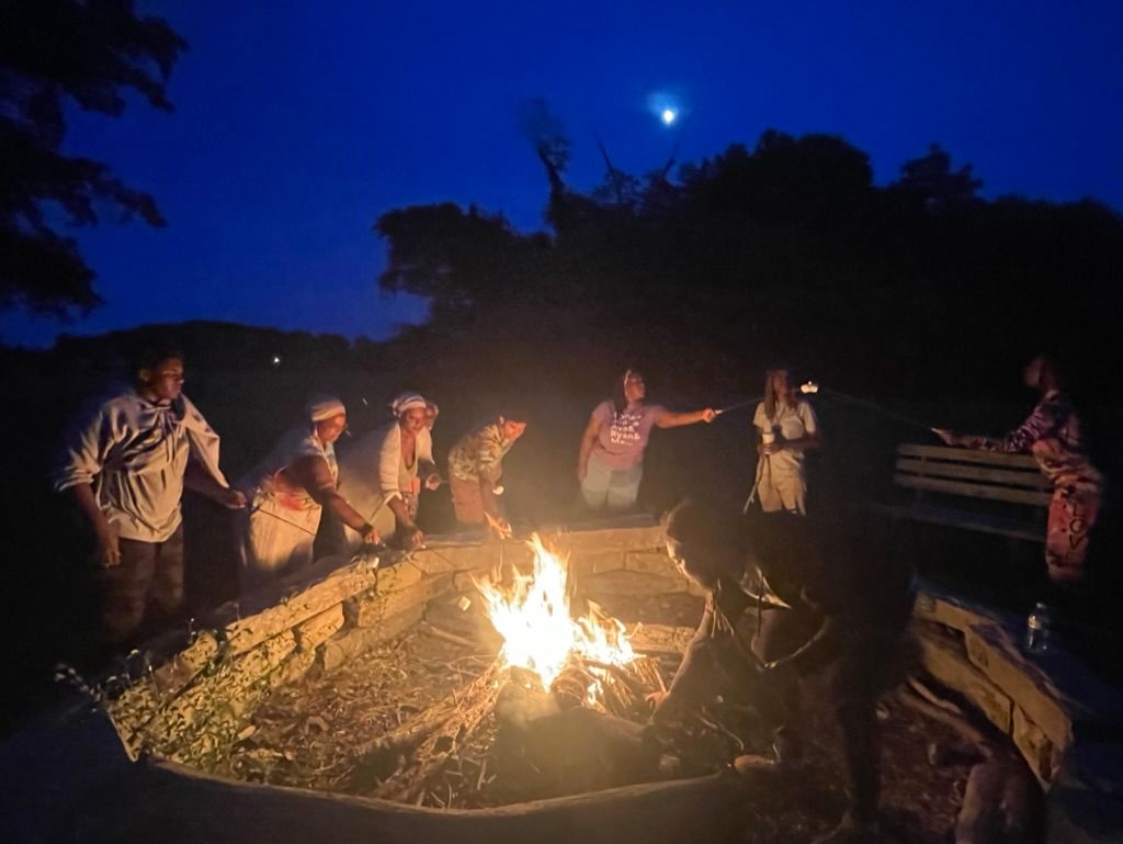 Eight people stand around a campfire at night under a deep blue sky