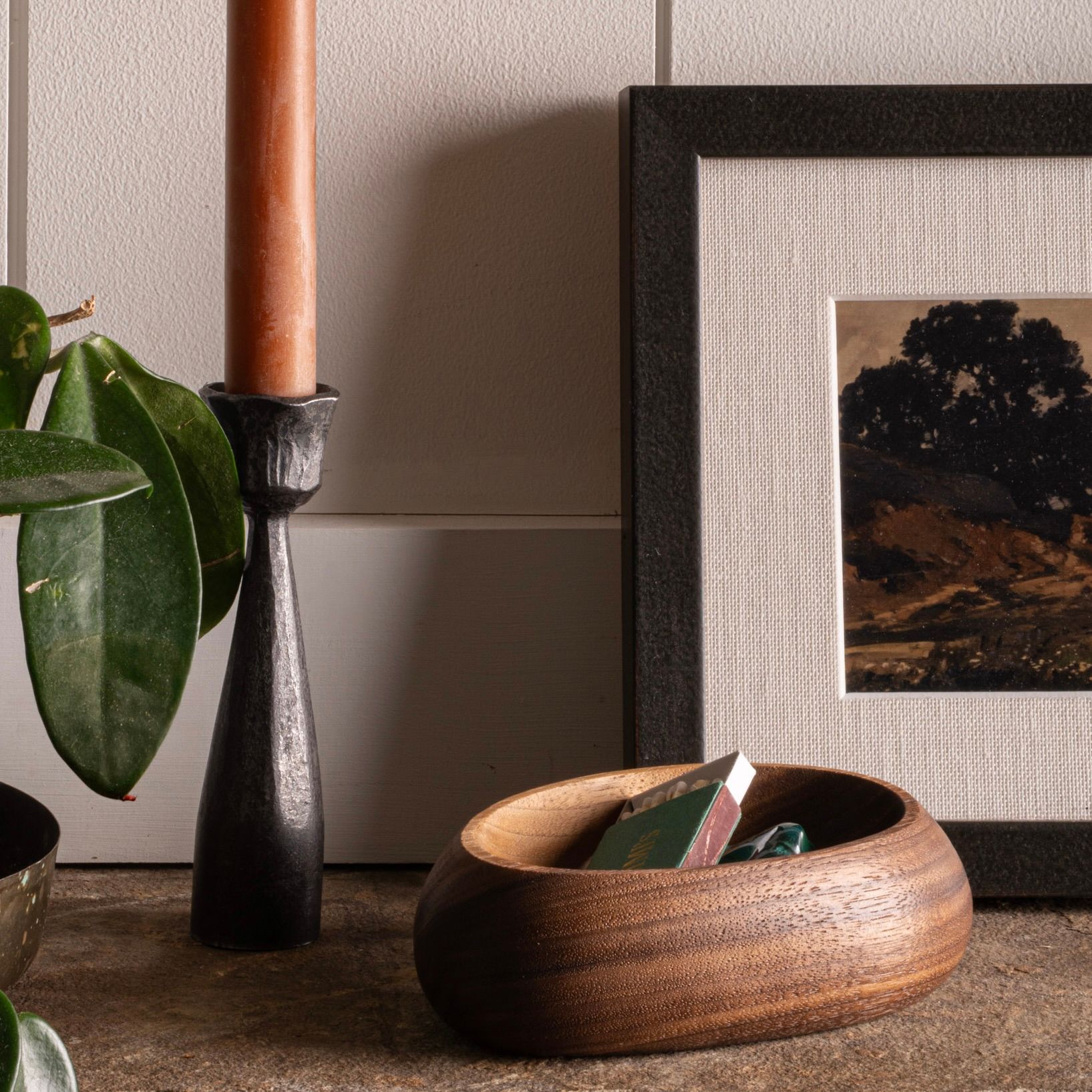 Wooden bowl with matchbox, black iron candlestick with taper candle, framed art, and plant on a mantle