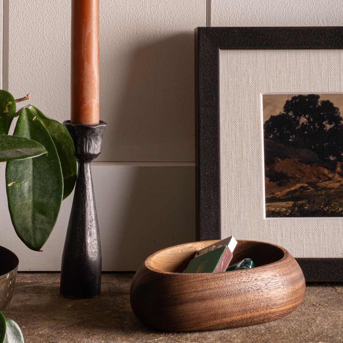 Wooden bowl with matchbox, black iron candlestick with taper candle, framed art, and plant on a mantle