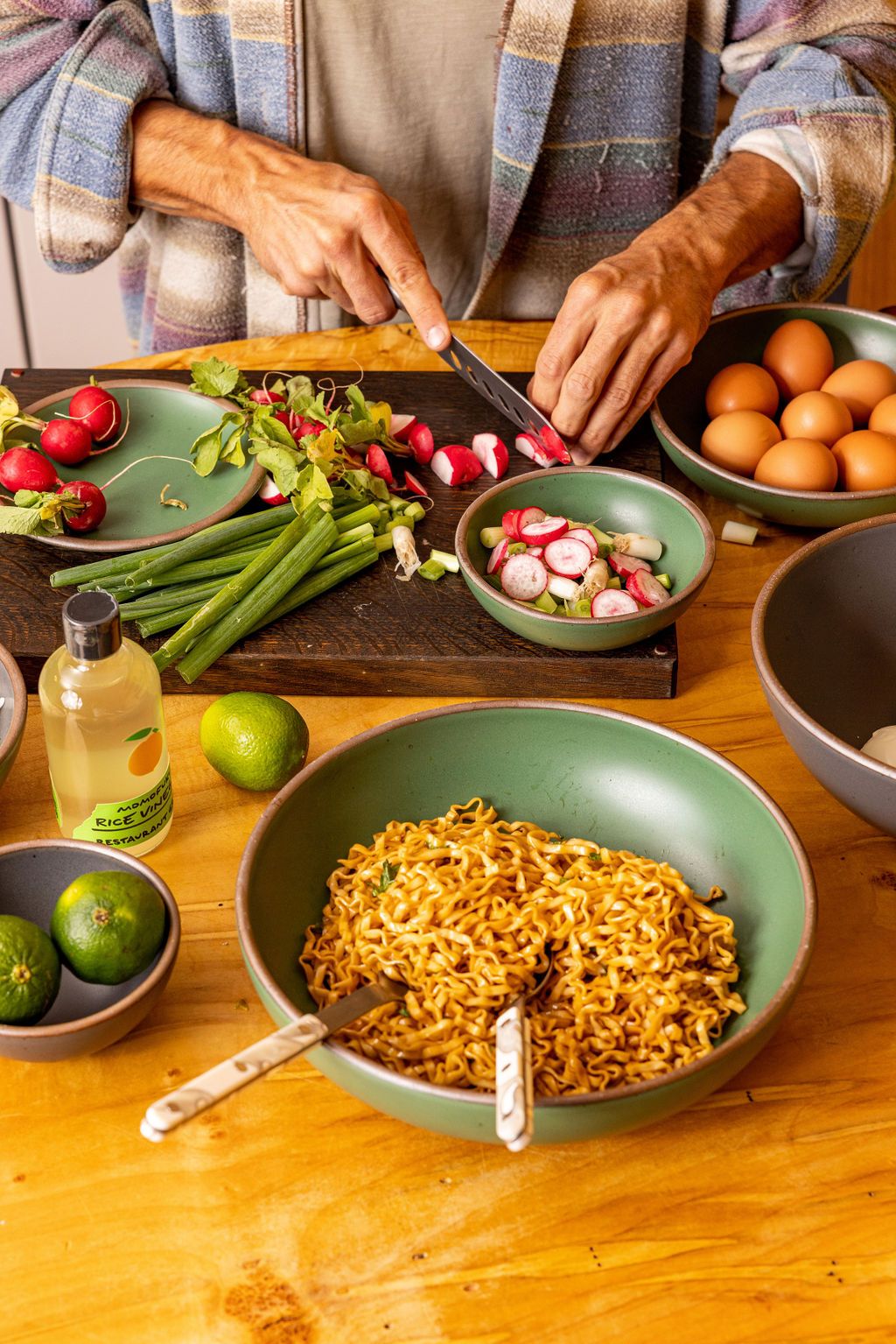 Image of someone chopping radishes and other vegetables on a counter with a green serving bowl of ramen in the forefront