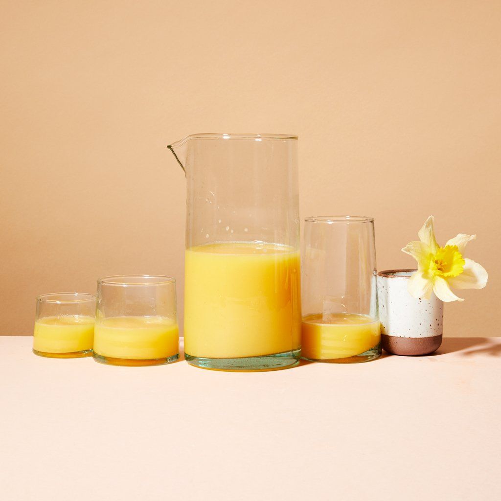 A glass cylindrical pitcher half full of orange juice surrounded by three matching glasses in different sizes and an East Fork Toddler Cup in Eggshell with a daffodil sticking out the top