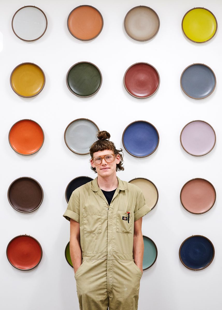 Kyle is wearing a jumpsuit and glasses with colorful ceramic platters displayed on the wall behind them.