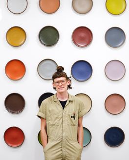 Kyle is wearing a jumpsuit and glasses with colorful ceramic platters displayed on the wall behind them.