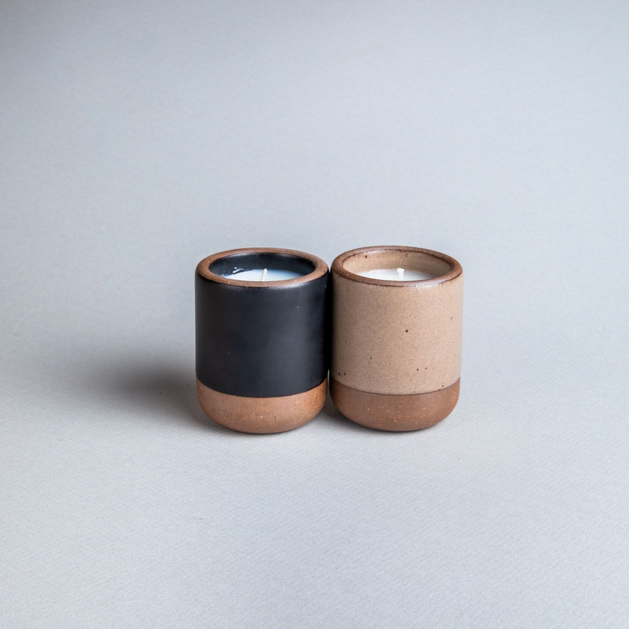 Two small ceramic vessels in dark charcoal and warm pale brown colors with candles inside each.