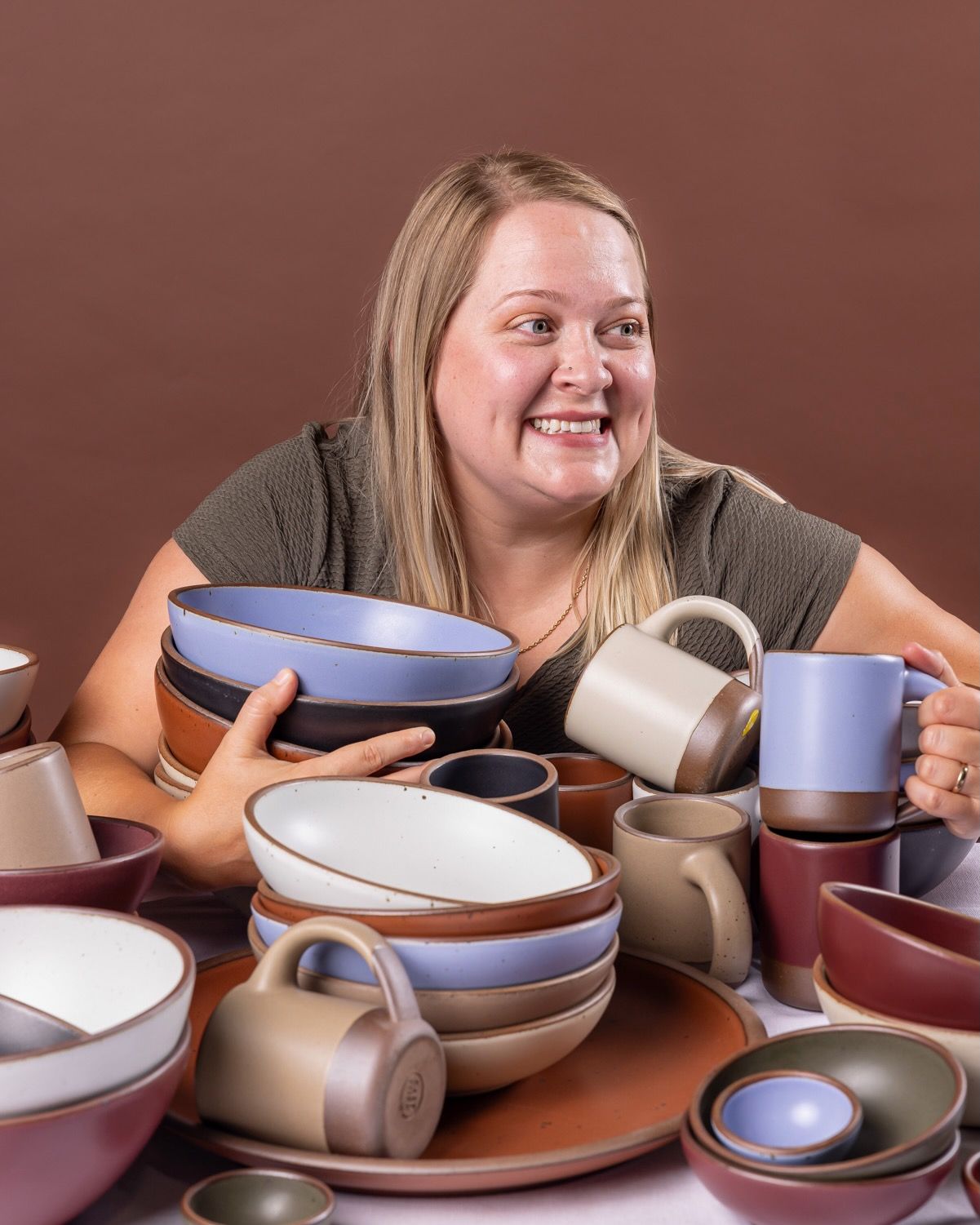 A woman holds onto a large stack of ceramic bowls, plates, and mugs all stacked on each other and smiles to the right.