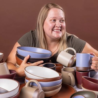 A woman holds onto a large stack of ceramic bowls, plates, and mugs all stacked on each other and smiles to the right.