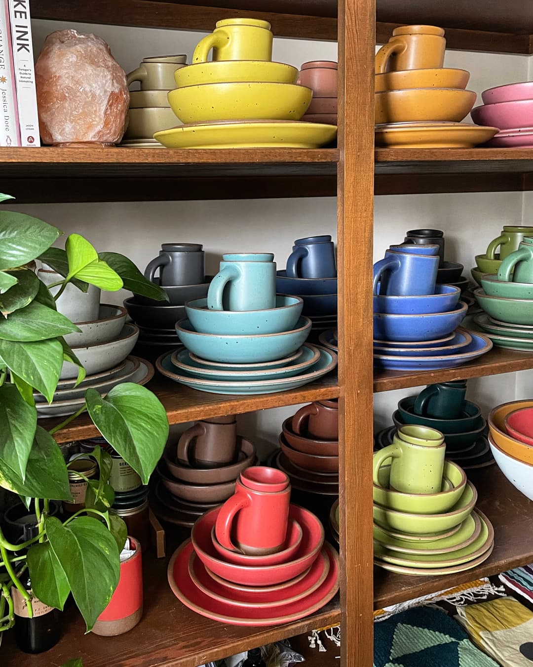On a shelf, are stacks of plates, bowls, and a mug - each in a different color of the rainbow.