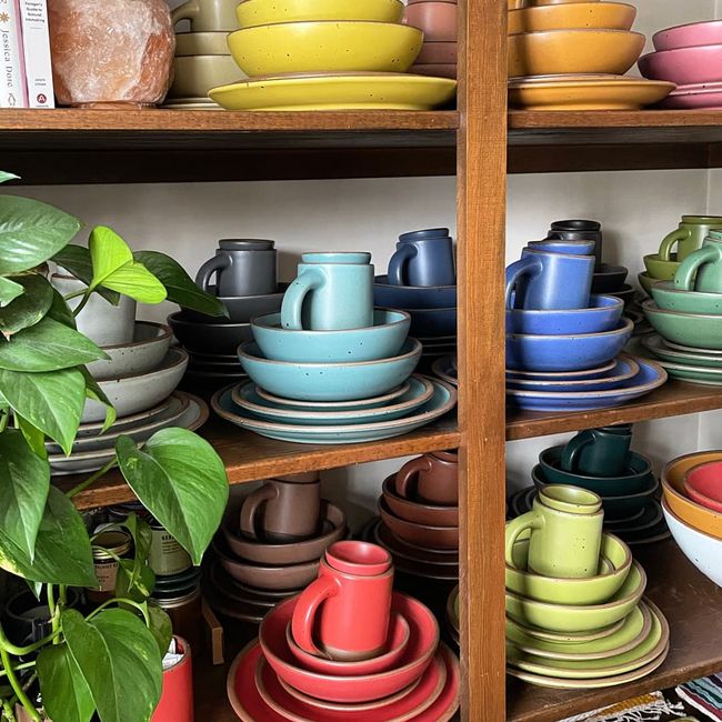 On a shelf, are stacks of plates, bowls, and a mug - each in a different color of the rainbow.