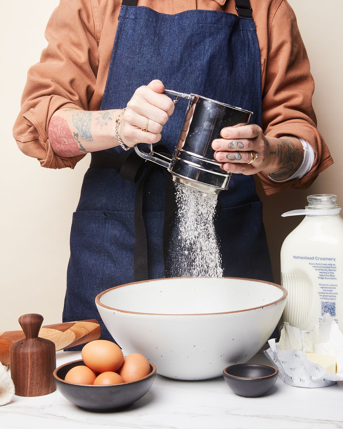 A person wearing a denim apron and holding a flour sifter over a mixing bowl. Surrounded are a biscuit cutter, a bowl with eggs, a carton of milk, opened stick of butter, and a bitty bowl.