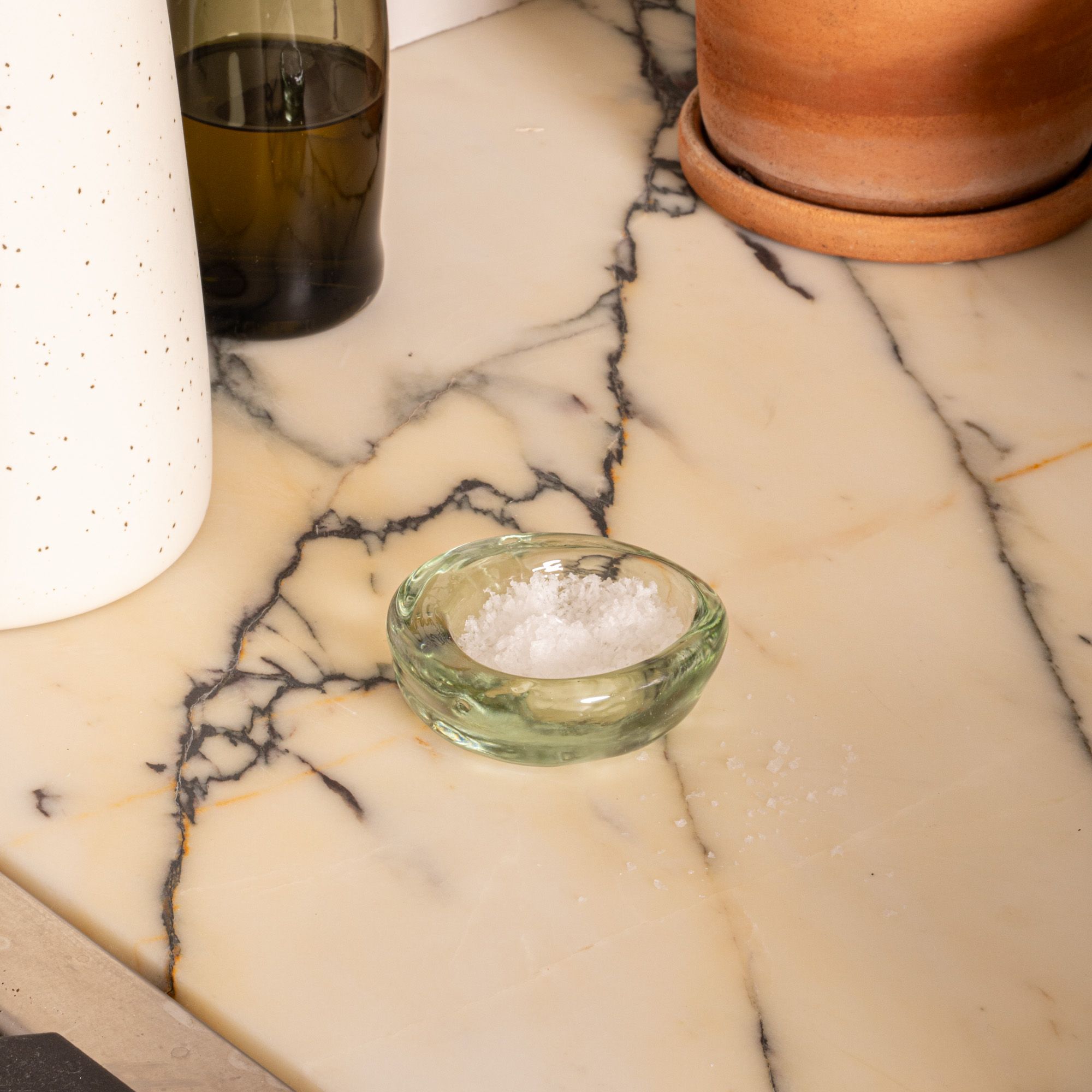 A small recycled glass bowl filled with flaky salt on a marble countertop near ceramic and glass containers.