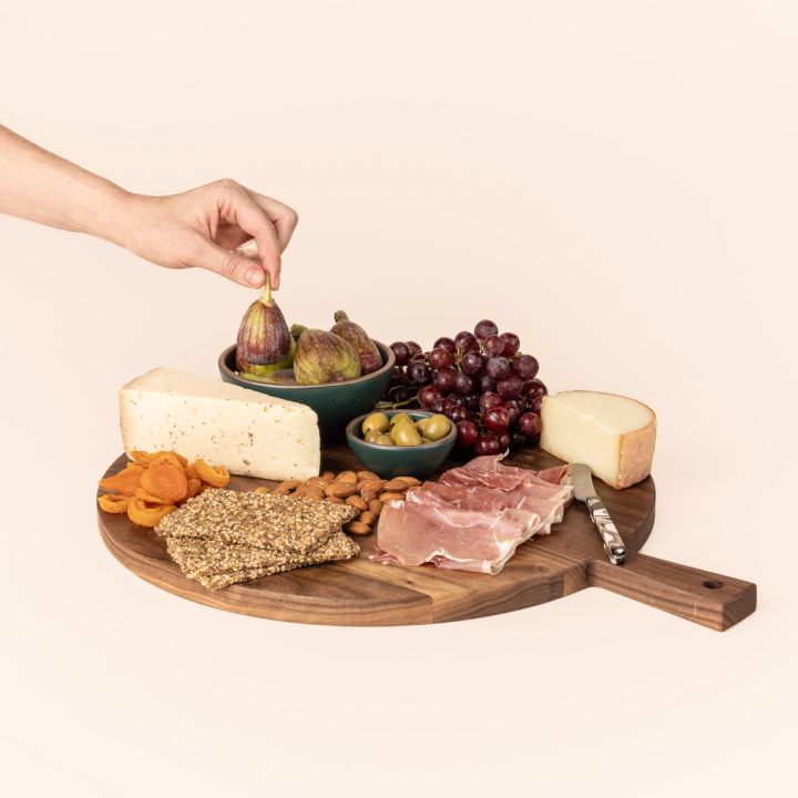 A hand grabbing food from a charcuterie spread on a round wooden serving board. The board has a short rectangular handle with a hole for hanging.