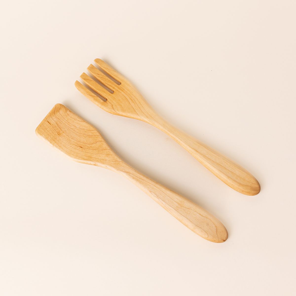 Two long maple wood serving utensils. The left utensil has a shape like a spatula, the right has a shape like a fork.