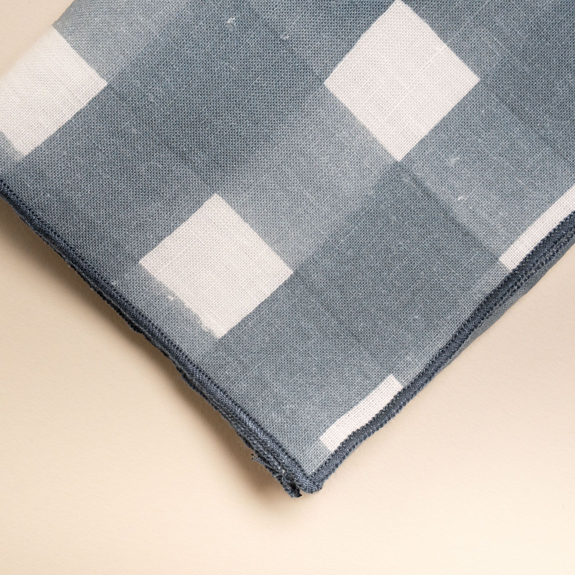 A closeup of the corner of a linen napkin in a blue-grey crosshatch pattern with navy serged edges