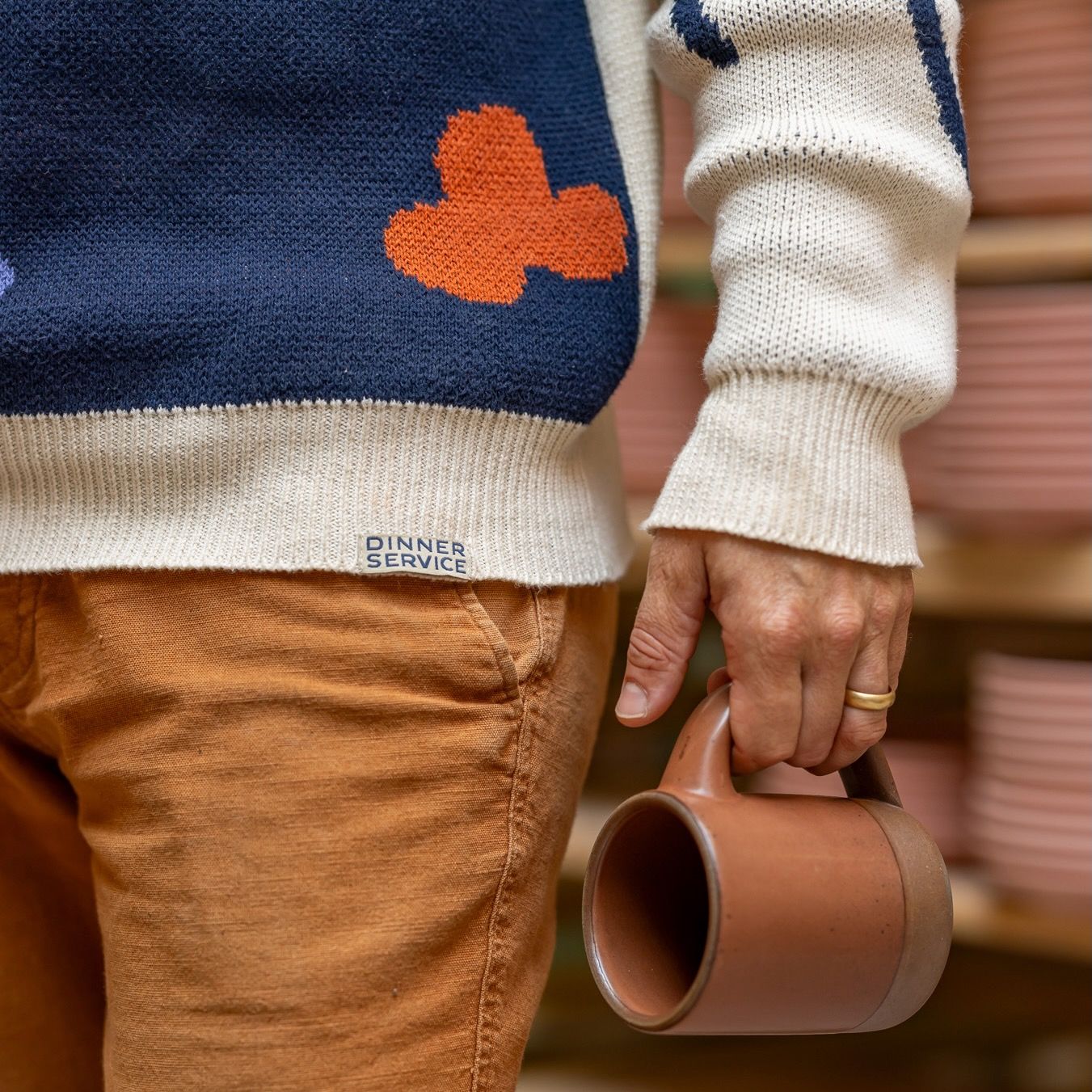 A closeup of the workshop sweater showing off the ribbed hem and cuff, with a hand holding a terracotta glazed mug.