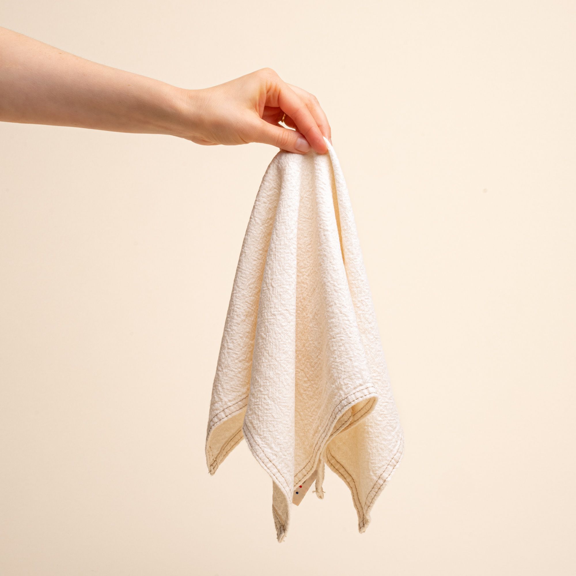 A hand holds the center of a cream linen napkin with sewn edge