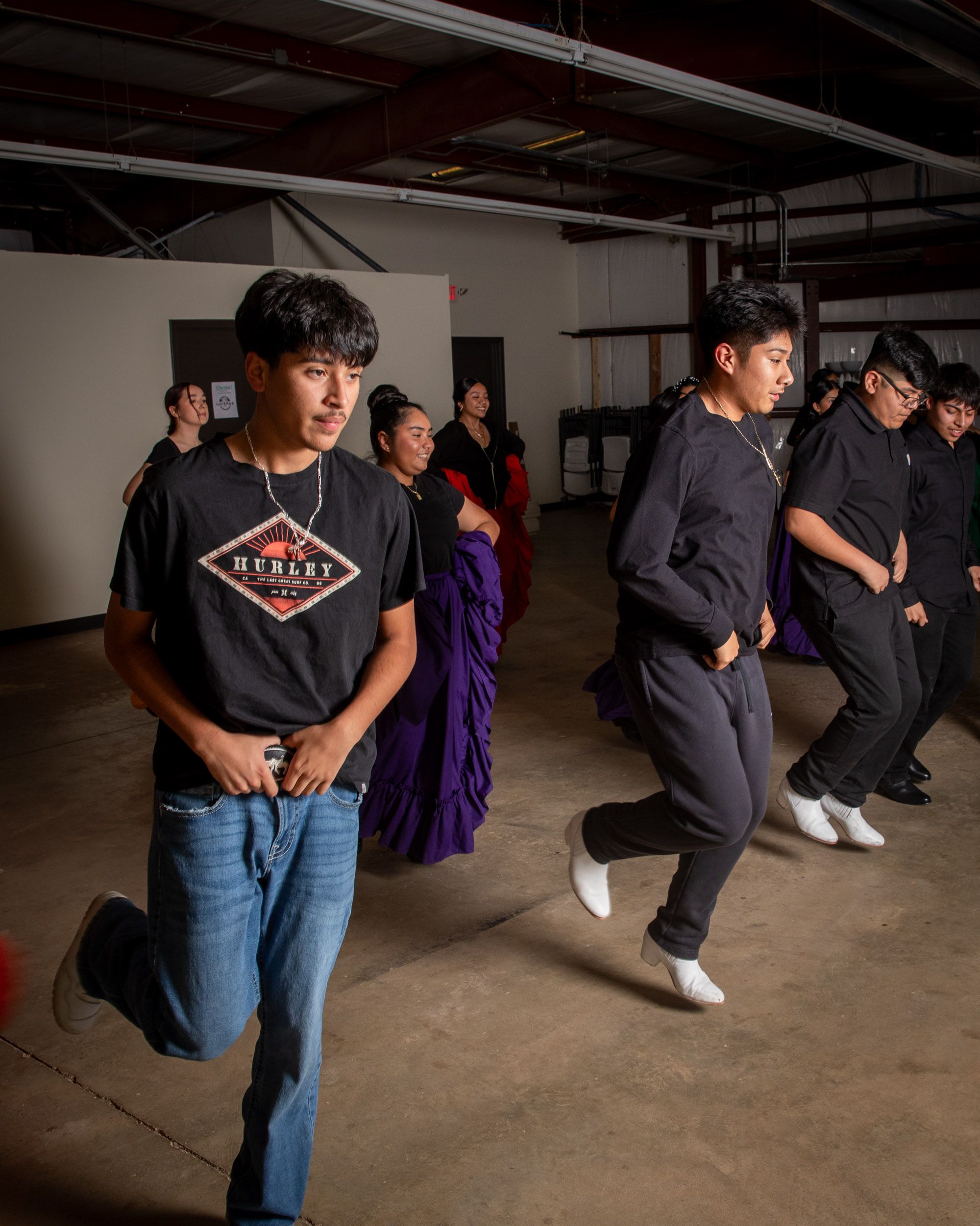 In a rehearsal space, there are young boys in casual clothes dancing in a row and holding their belt buckles.