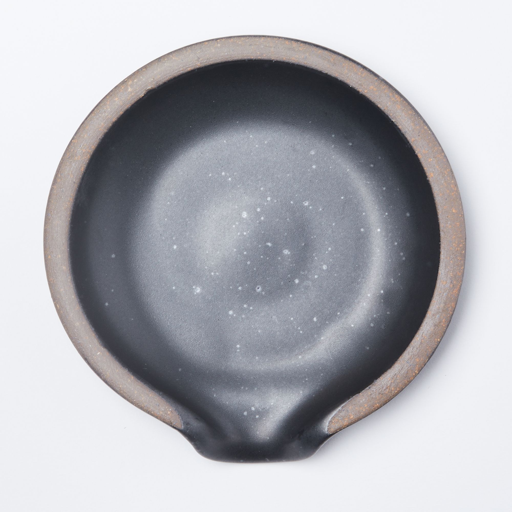 A circular ceramic spoon rest in a graphite black color featuring iron speckles and unglazed ridge rim