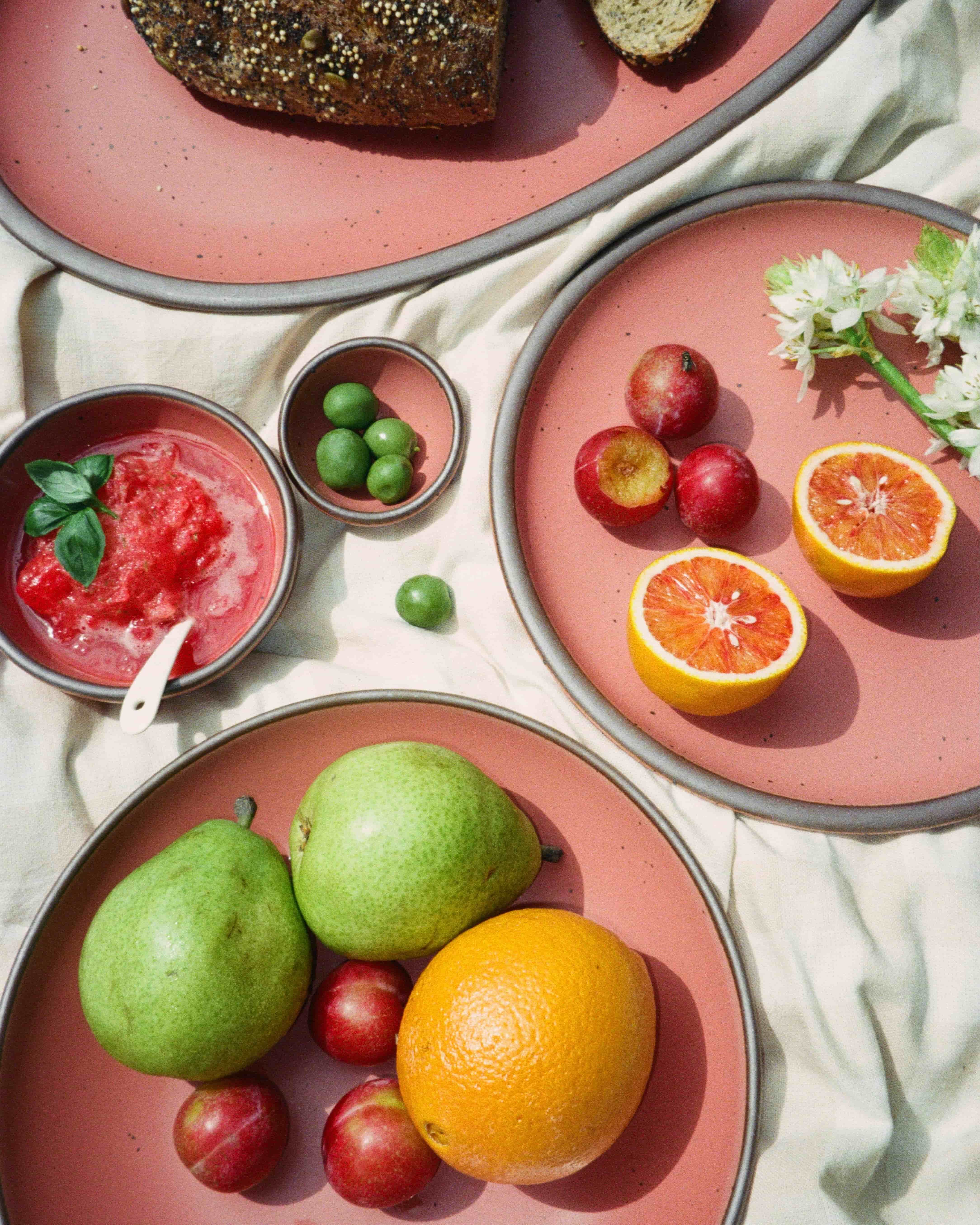 Assorted fruits, bread, and spread served on watermelon pink ceramic plates laid out on a light cloth for a picnic-style setup.