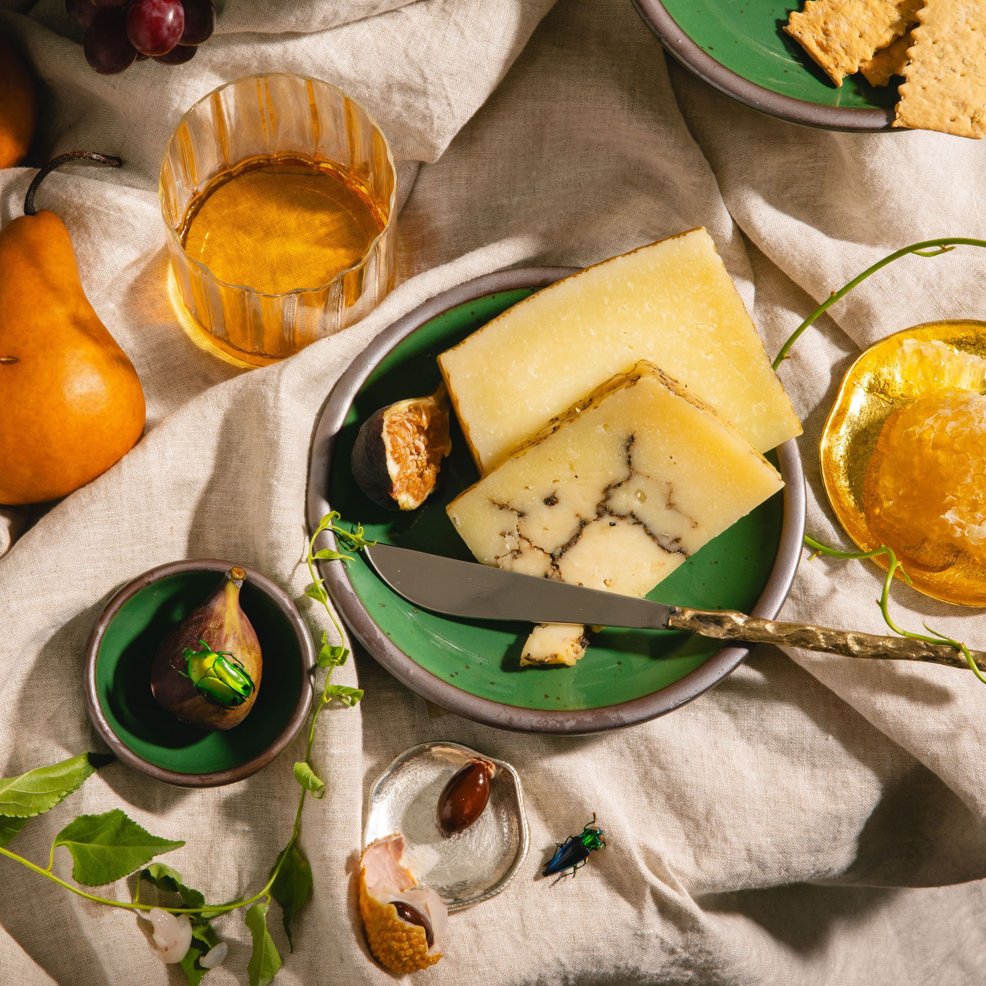 Glossy emerald green plate with truffle cheese, figs, crackers, honeycomb, and a glass of amber drink on linen.