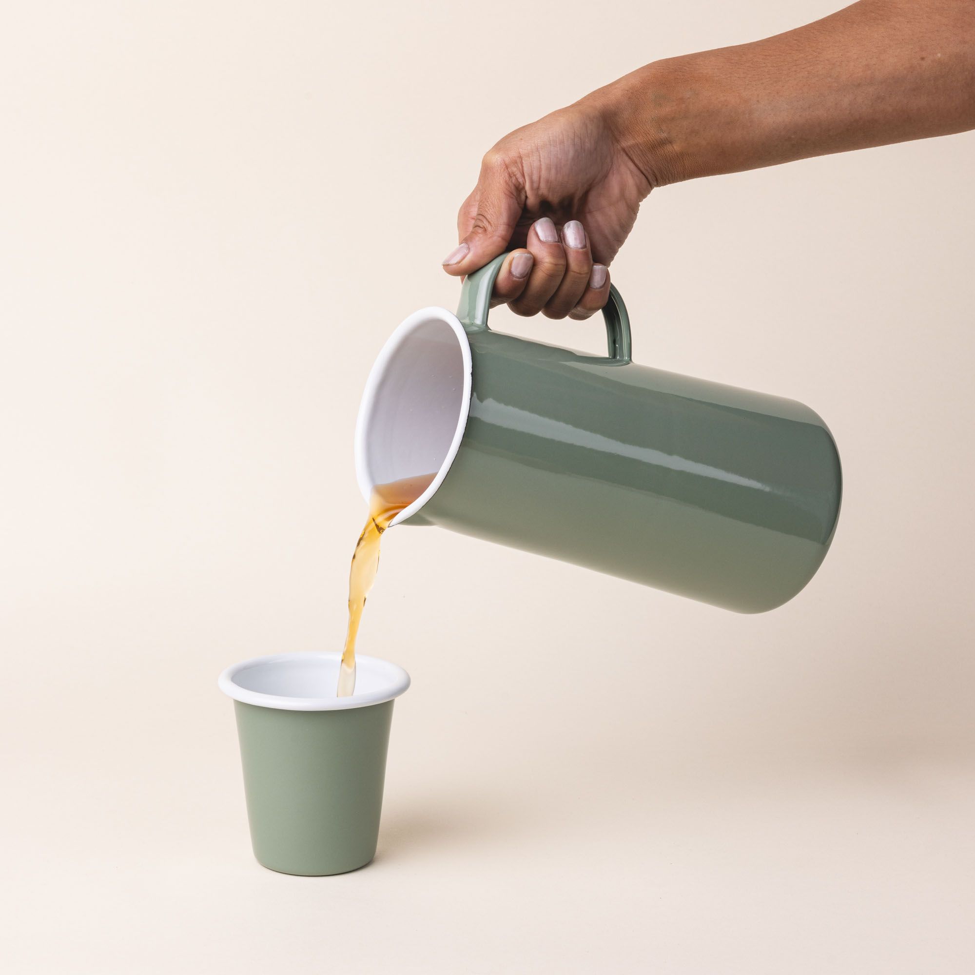 An enamel pitcher pours tea into a short enamel cup with a sage green exterior and white interior