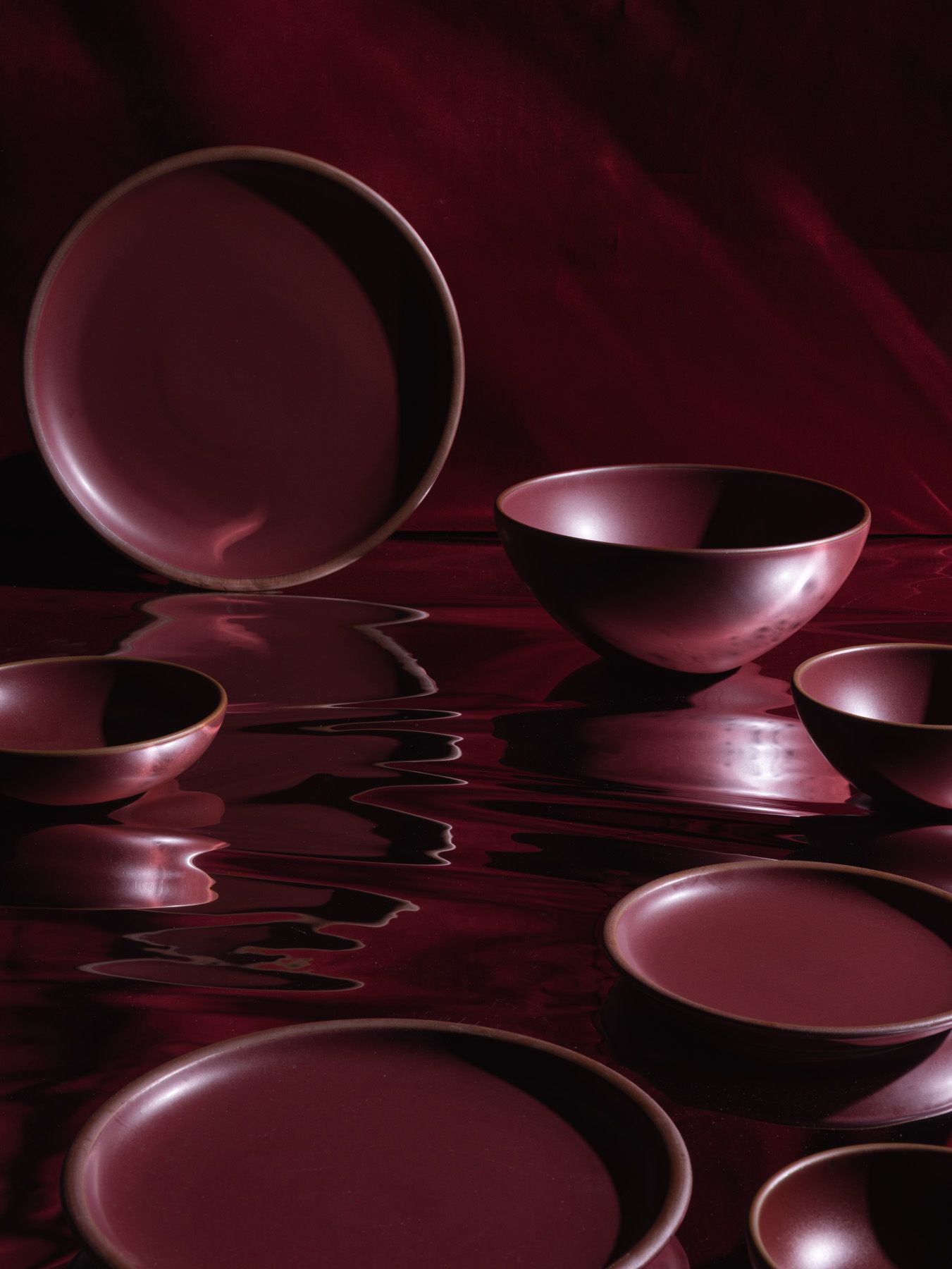 A moody artistic setting of dark plum ceramic dinnerware arranged on a mirrored surface that mimics a dark sea. 