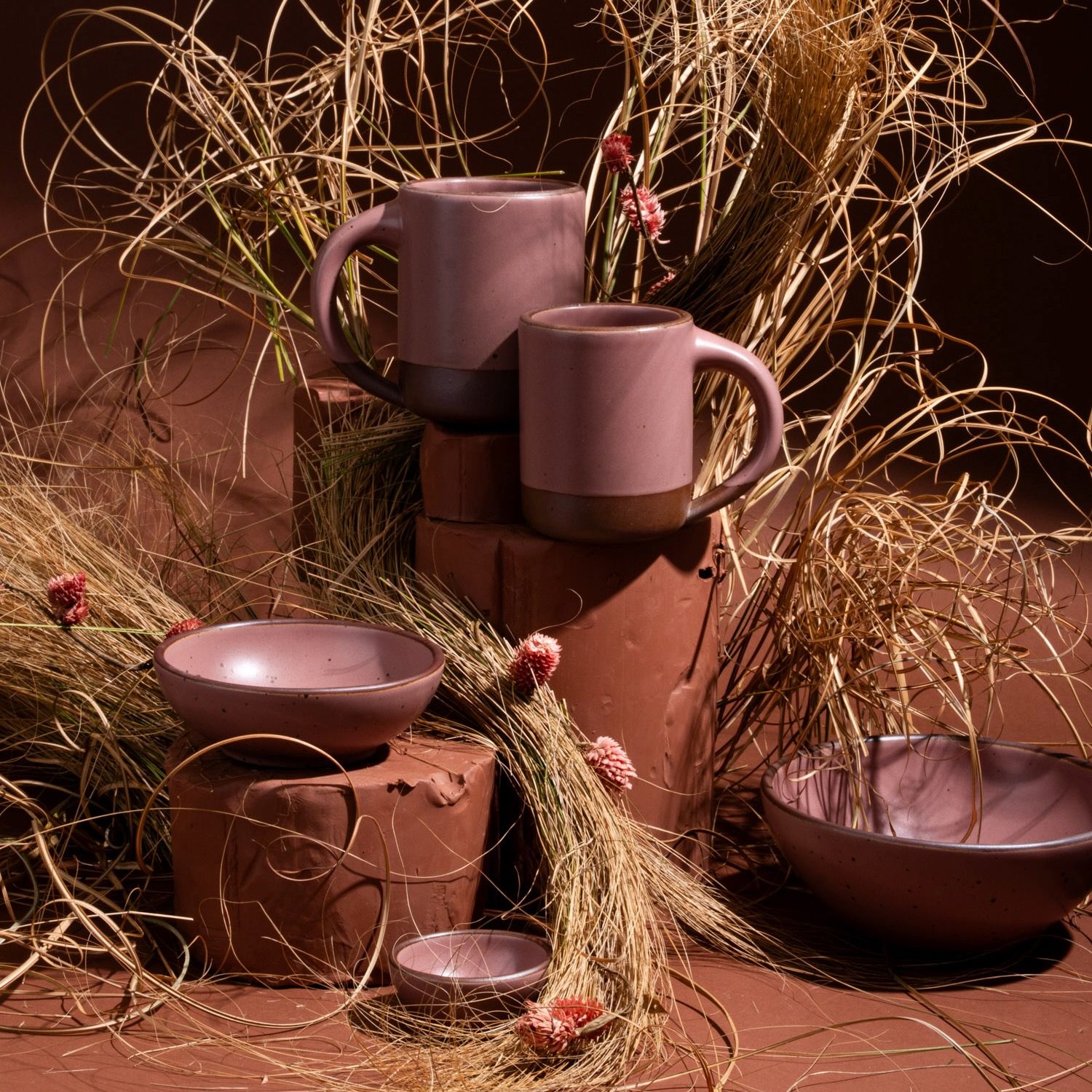 Dusty mauve pink mugs and bowls artfully arranged with clay with dried grass and flowers