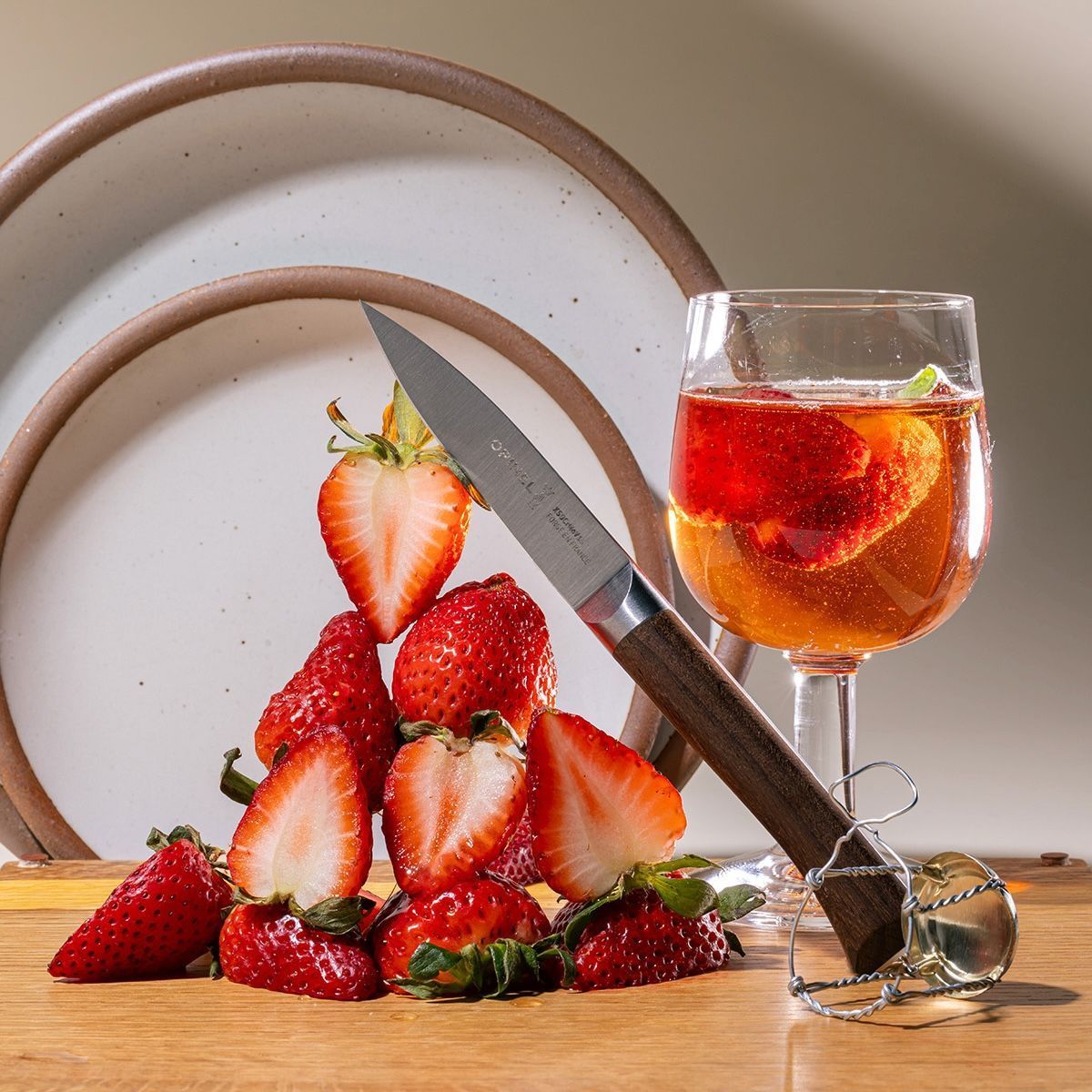 A small paring knife is leaning against a tower of sliced strawberries and sitting on a cutting board. Nearby are a stack of white ceramic plates in the background and a wine glass filled with a sparkling drink and sliced strawberries. The knife has a sharp steel blade and a beveled dark wood handle