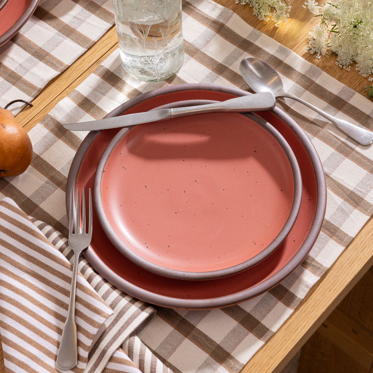 Stacked watermelon pink stoneware plates with silverware on a checked table runner and striped napkin, next to a glass of water.