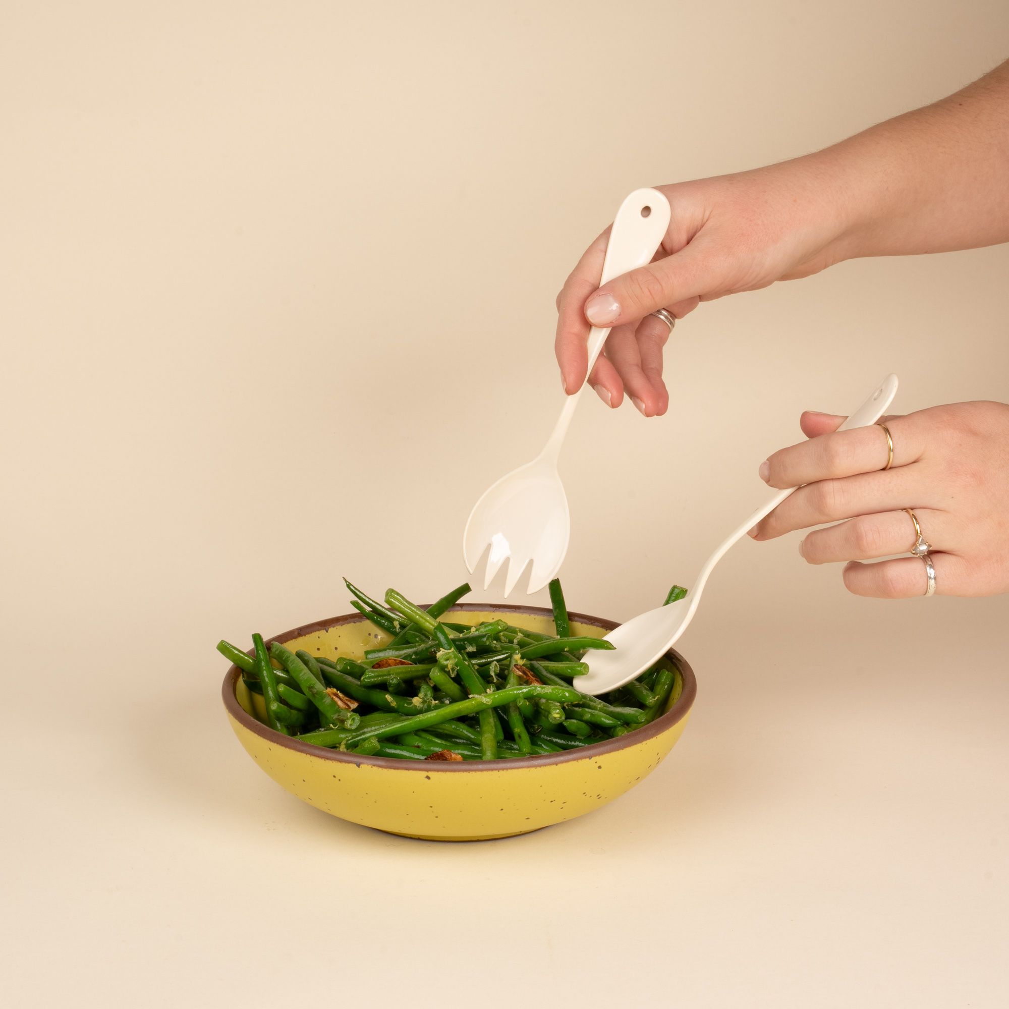 A hand holds a serving fork and spoon set made of white enamel above a bowl of green beans.