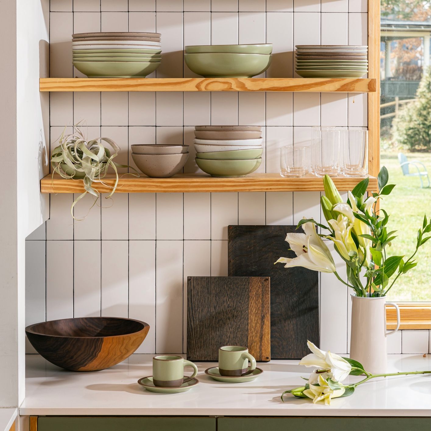 In a kitchen setting, there are floating shelves above a countertop filled with stacks of bowls and plates, simple drinking glasses, and an air plant. On the countertop is small mugs sitting on saucers, a large walnut bowl, 2 large dark cutting boards, and a water pitcher filled with flowers.