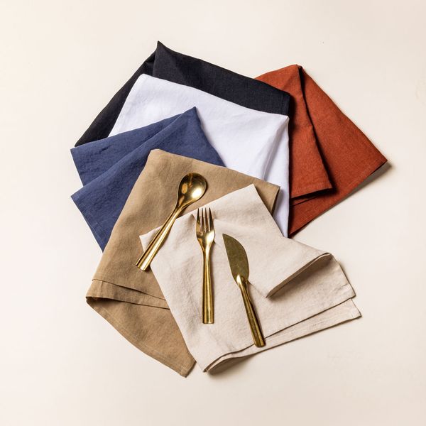 Six folded napkins arranged artfully in a stacked spiral with a brass spoon, fork, and knife laying on top. The napkins are in terracotta, black, white, navy, warm brown and off-white colors.