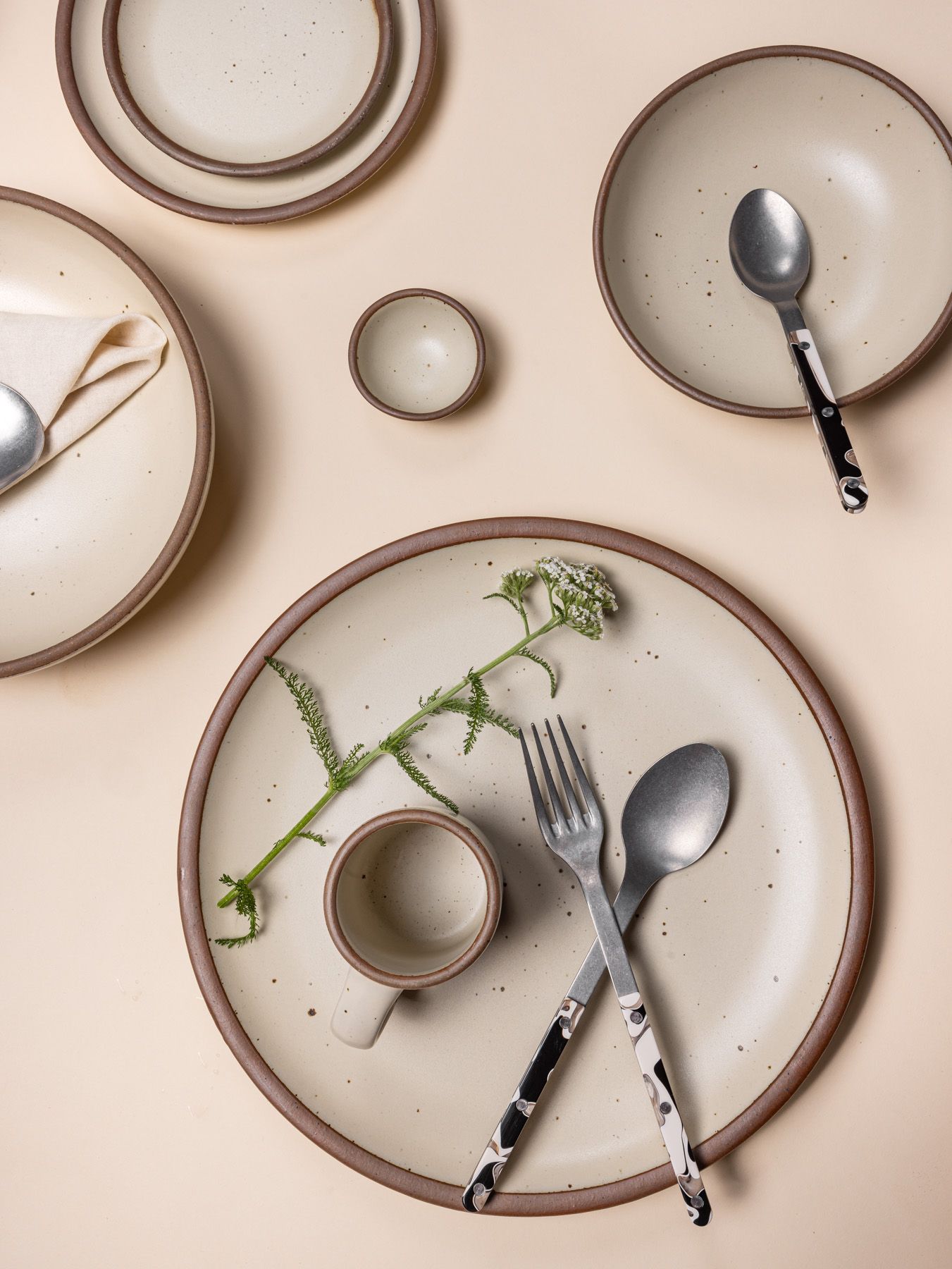 Overhead view of ceramic plates in various sizes in a neutral color with black marbled handled silverware and simple greenery