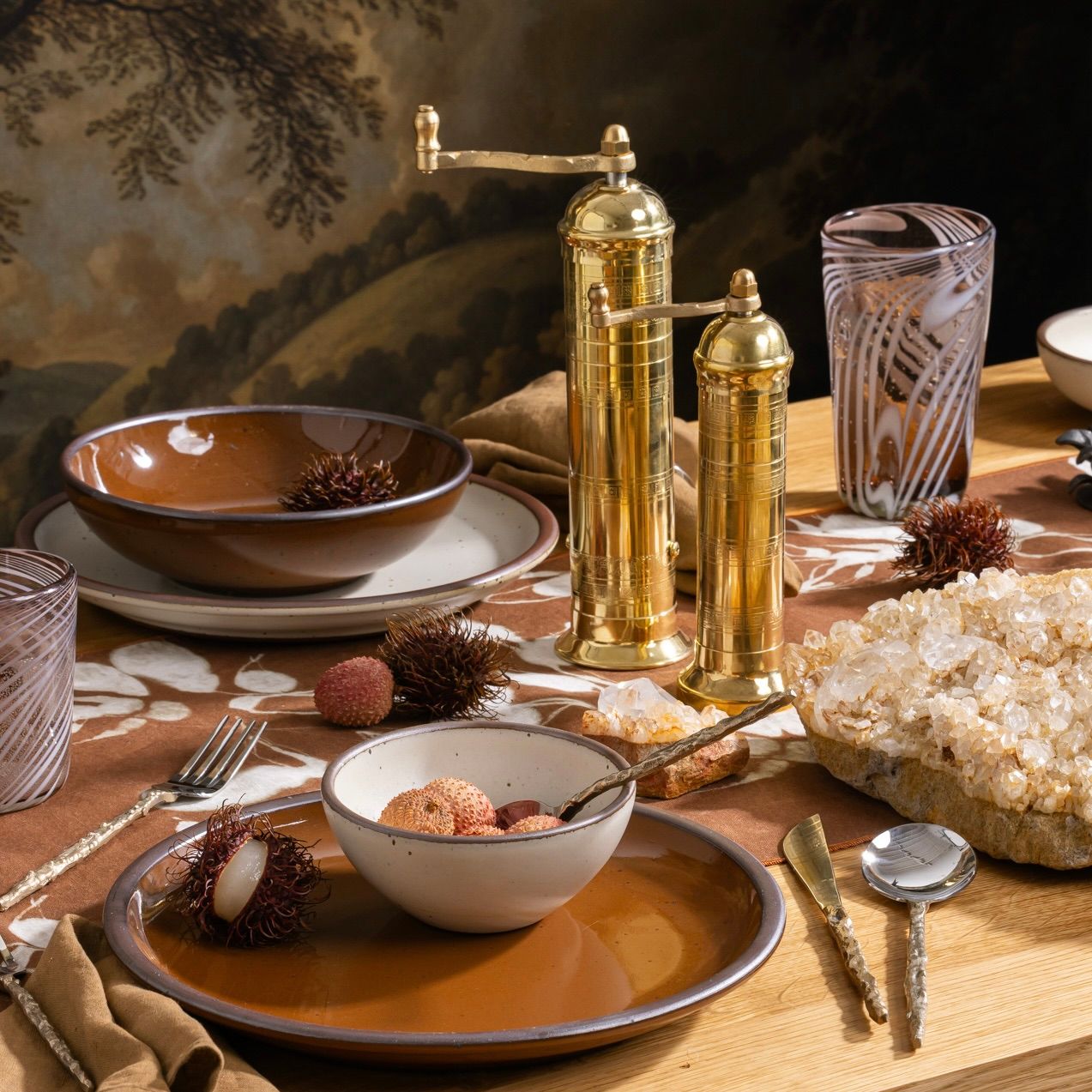 Brass salt and pepper mills on a set dining table with stoneware dishes, lychee fruit, crystal cluster, and patterned glassware.
