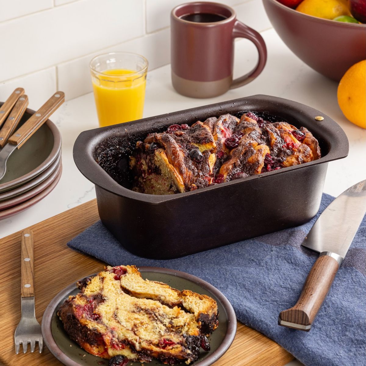 On the counter is an iron loaf pan filled with a cranberry loaf, slice on a cake plate, and some kitchen utensils.