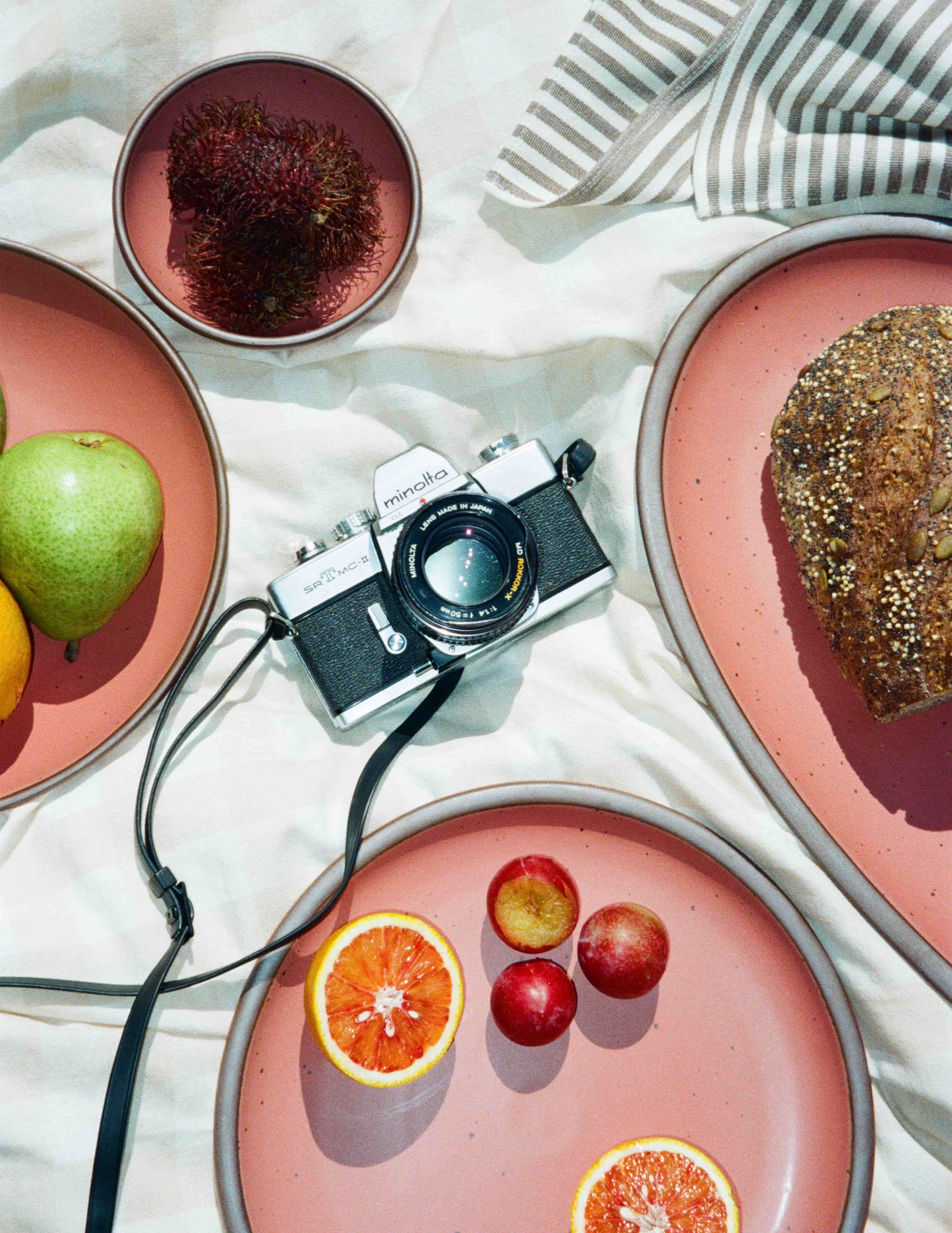 Minolta film camera surrounded by fruit and bread on watermelon pink ceramic plates over a white picnic cloth.